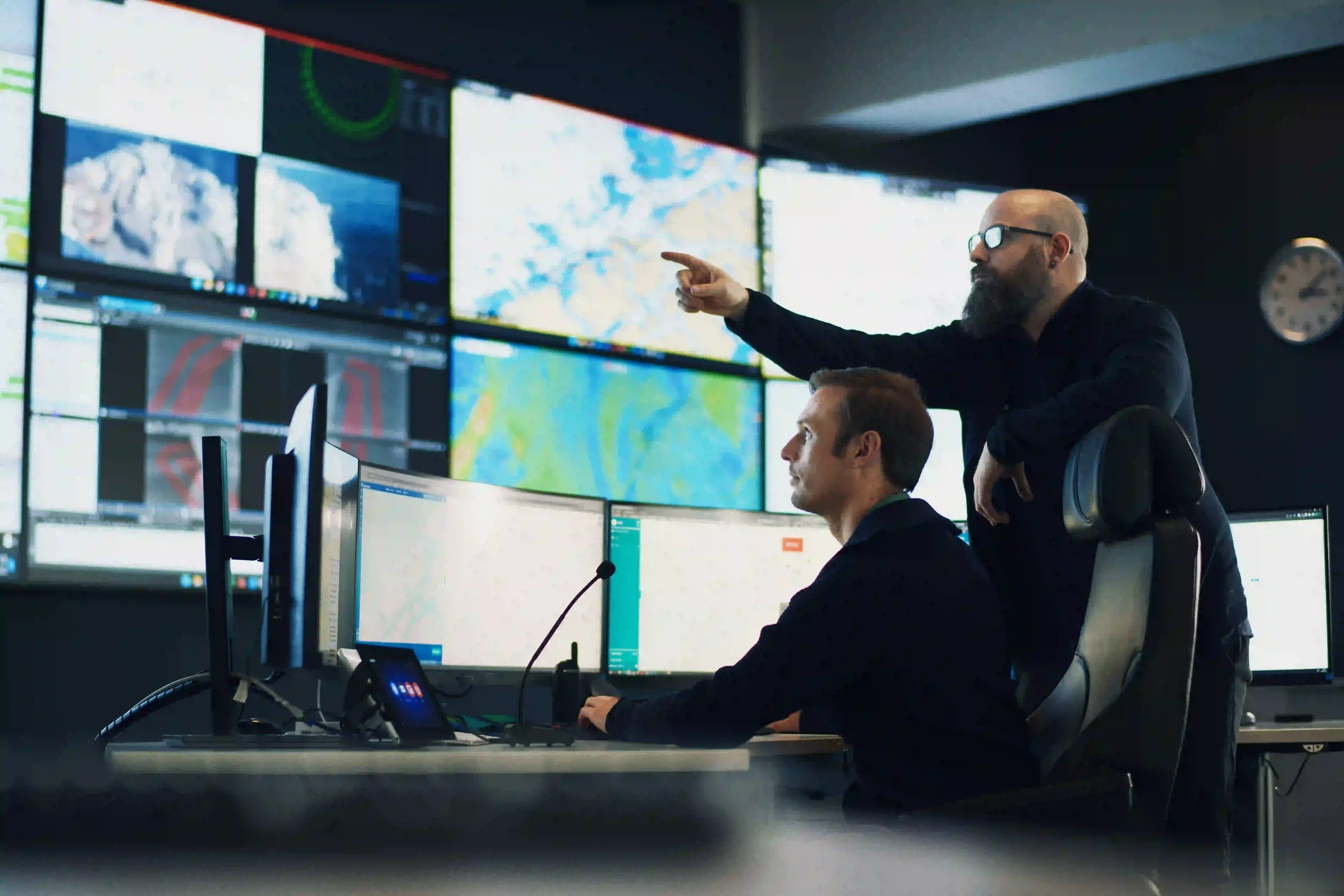 To personer i overvåkningssentralen Norseye Control, en person peker på flere skjermer med kart og data, mens den andre sitter foran flere dataskjermer.