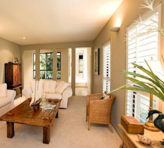Living room with beige sofas, wooden coffee table, wicker armchair, and large windows with white shutters.