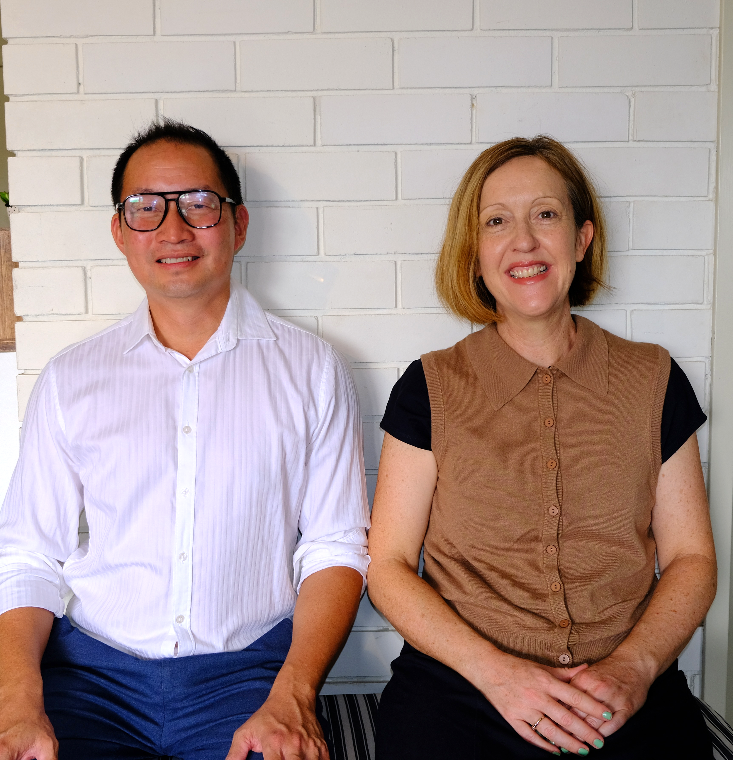 A man and a woman sitting side by side in front of a white brick wall, smiling at the camera.