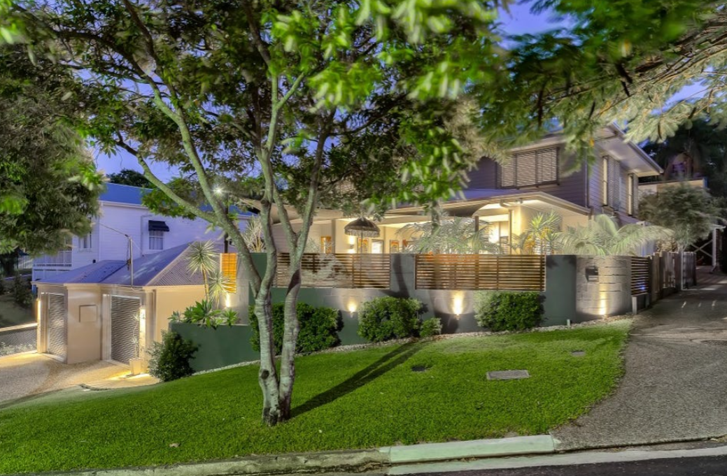 A contemporary house at night with exterior lighting, a wooden fence, a lush green lawn, trees, and an adjacent driveway.