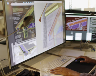 Multiple computer monitors displaying architectural or engineering design plans on a desk with a person working.
