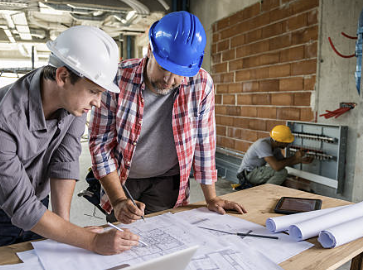 Two construction workers in hard hats discussing blueprints at a construction site, with another worker installing electrical panels in the background.