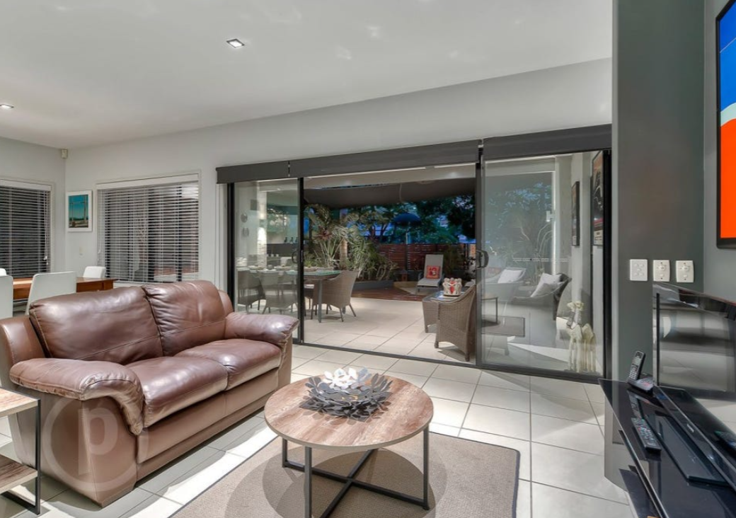 Living room with brown leather sofa, round wooden coffee table, and sliding glass door leading to outdoor patio with table and chairs.