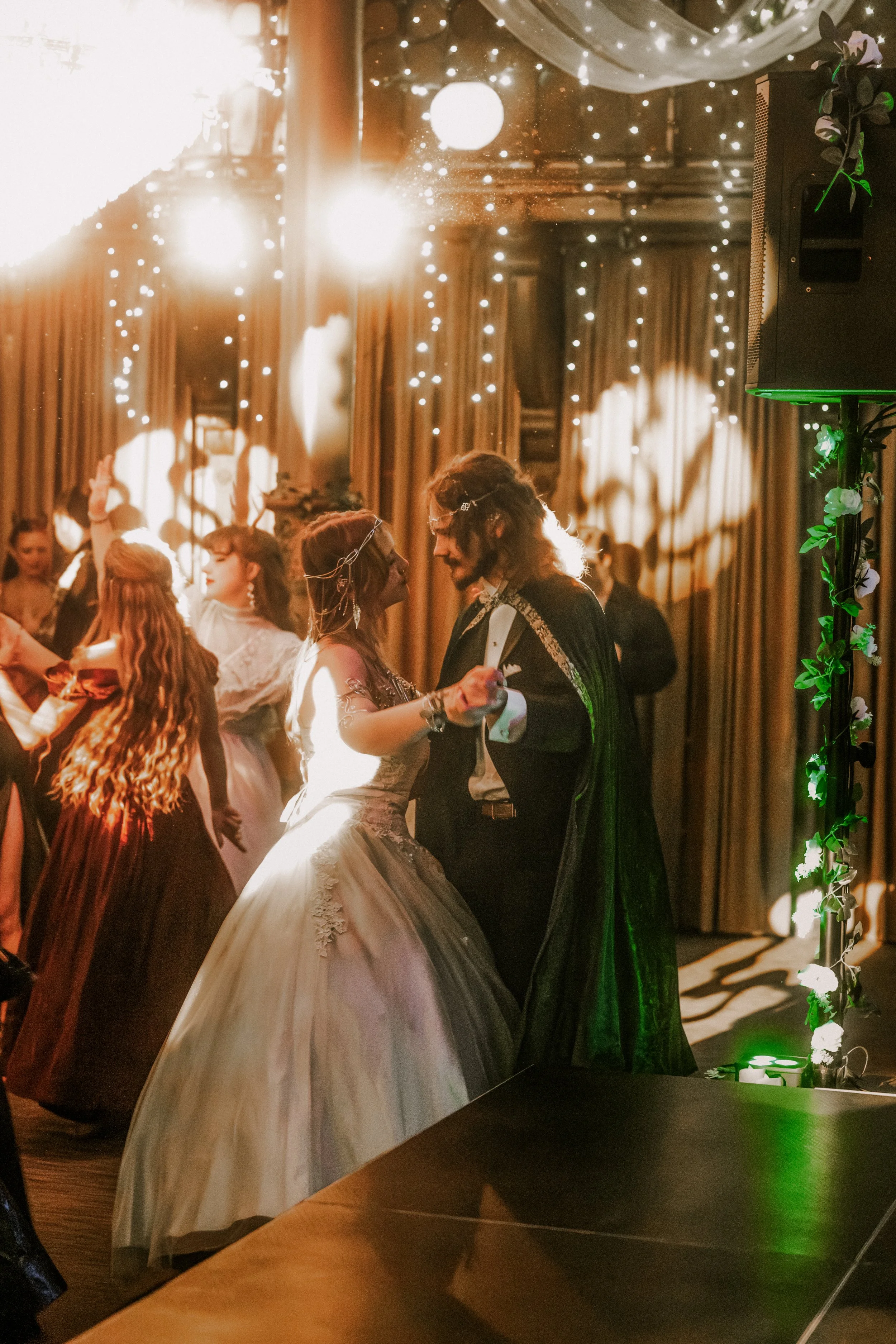 A couple dance during the fantasy ball as other guests dance in the background, with warm lighting, string lights, and decorated curtains.