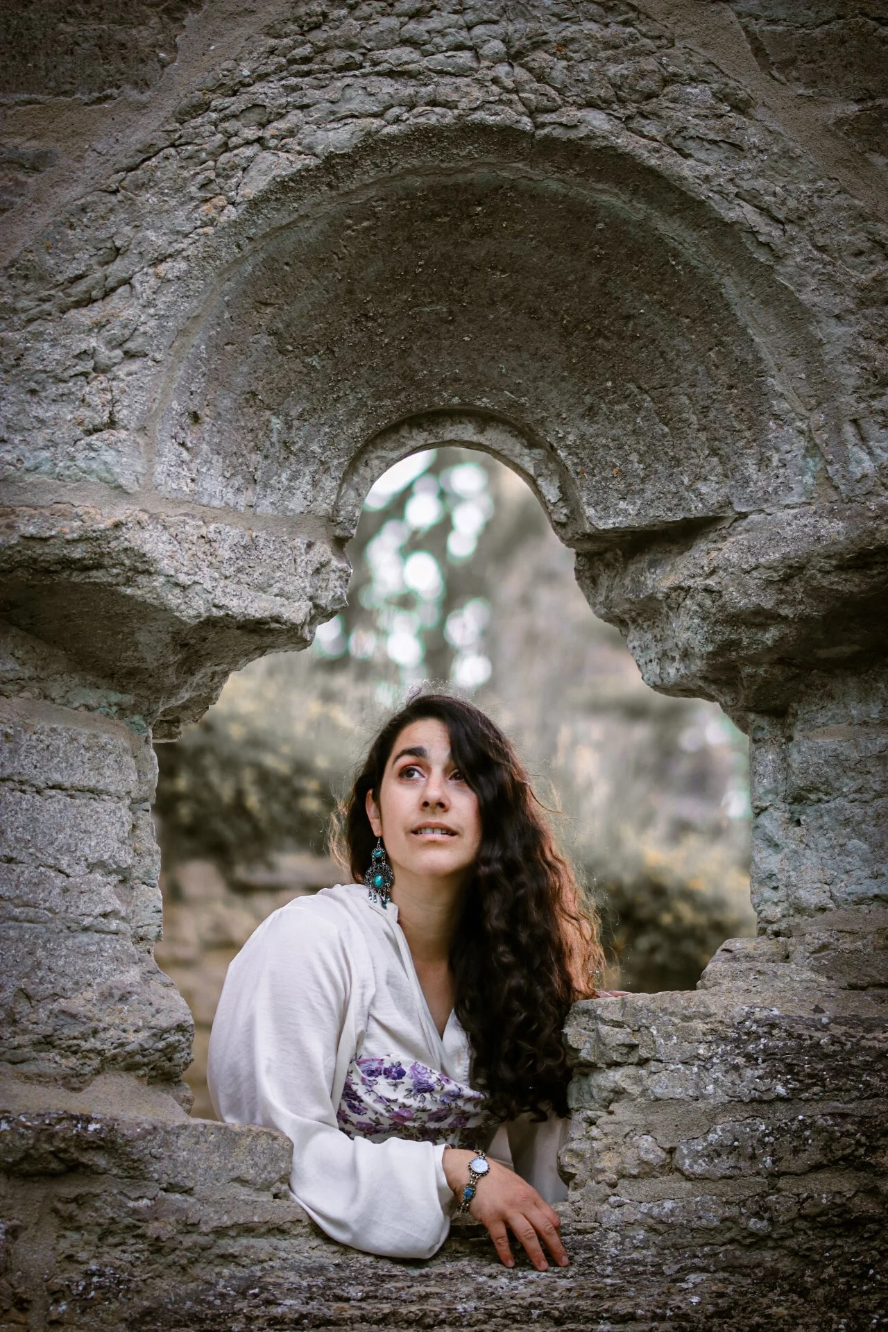 A woman with dark curly hair looks with longing out of a ruin window dressed in a flowy shirt and corset