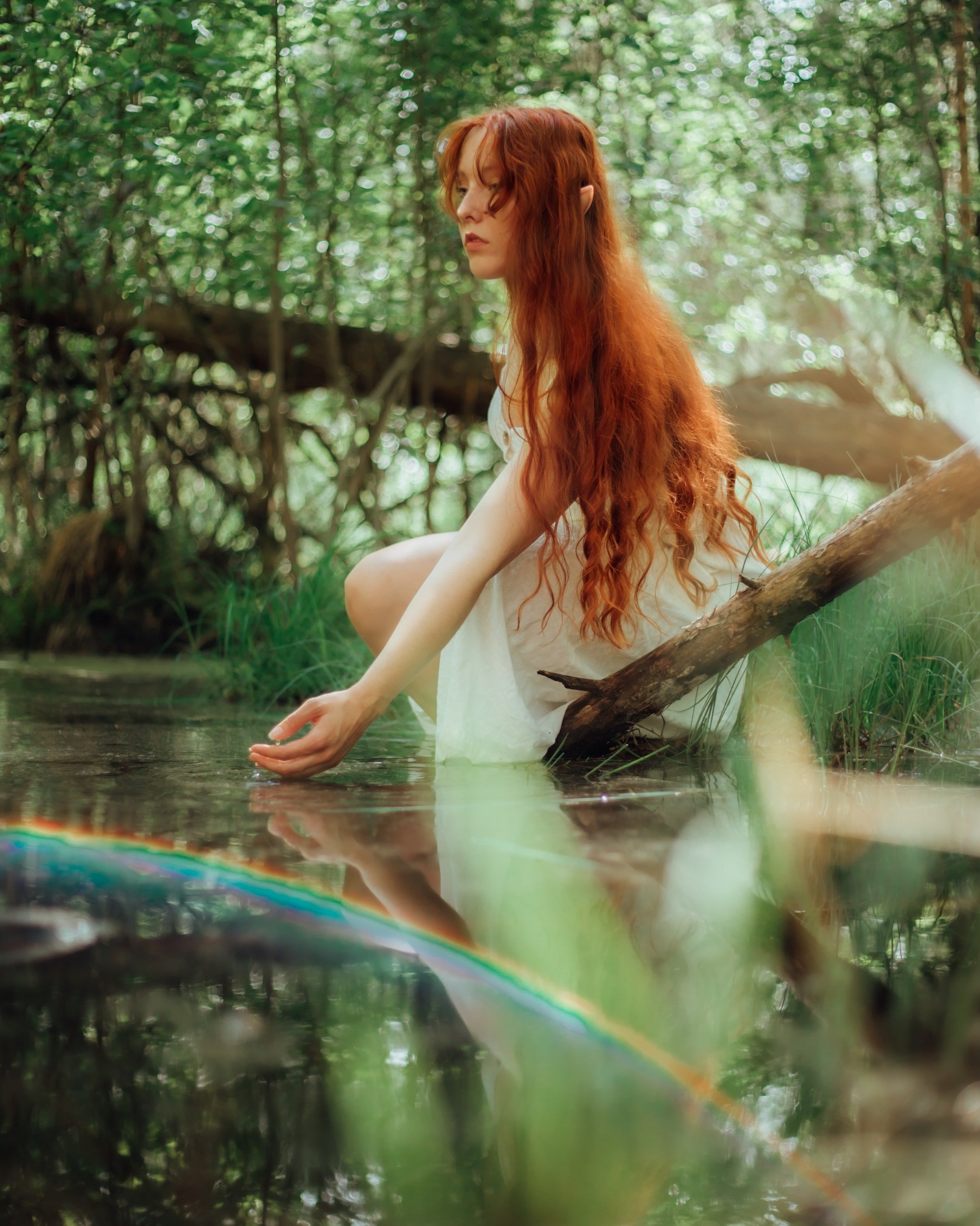 A woman with long red hair leans down to the water of a creek, surrounded by ancient woods with a rainbow reflected in the water