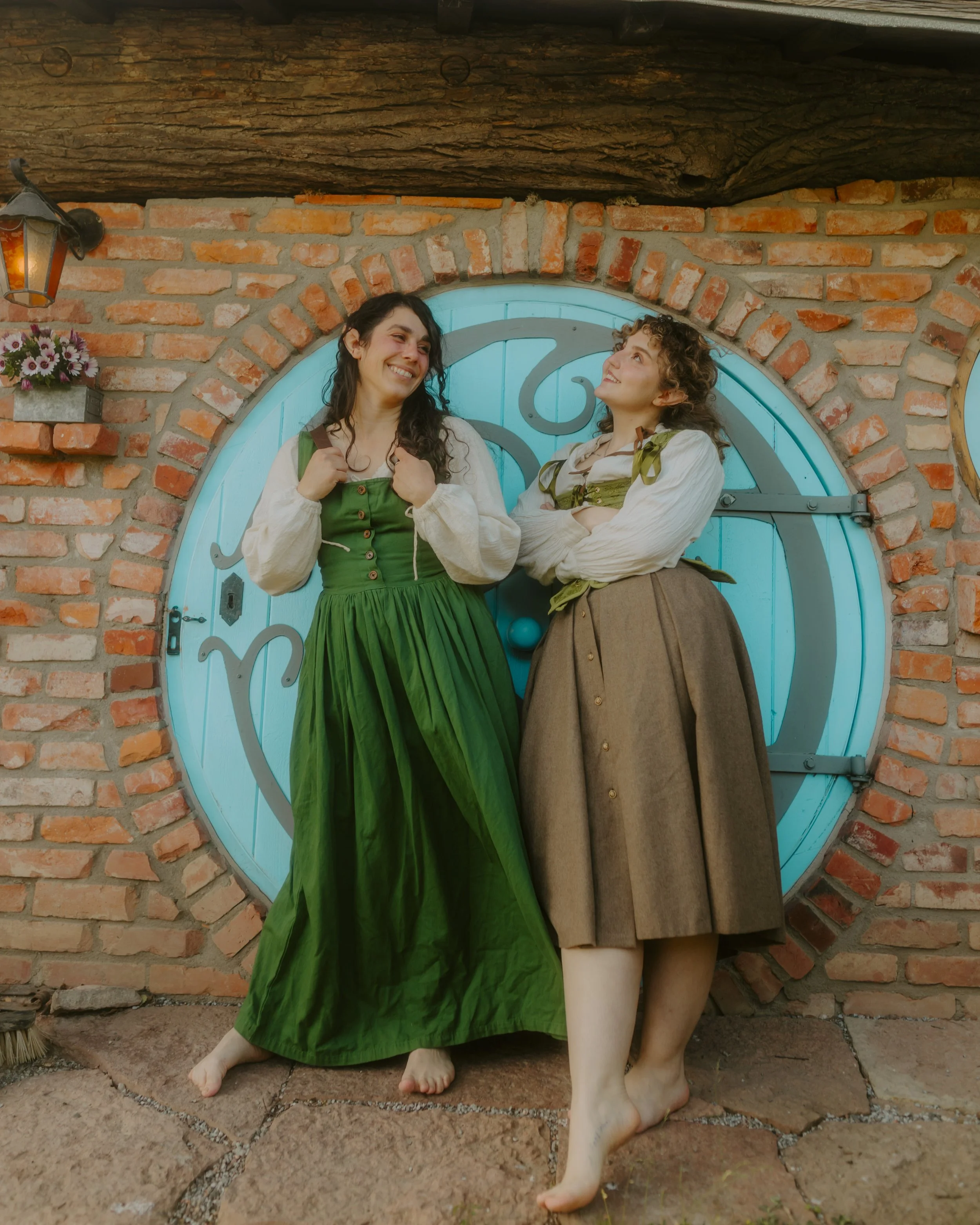 Two hobbit women standing in front of a round hobbit door smiling at each other.