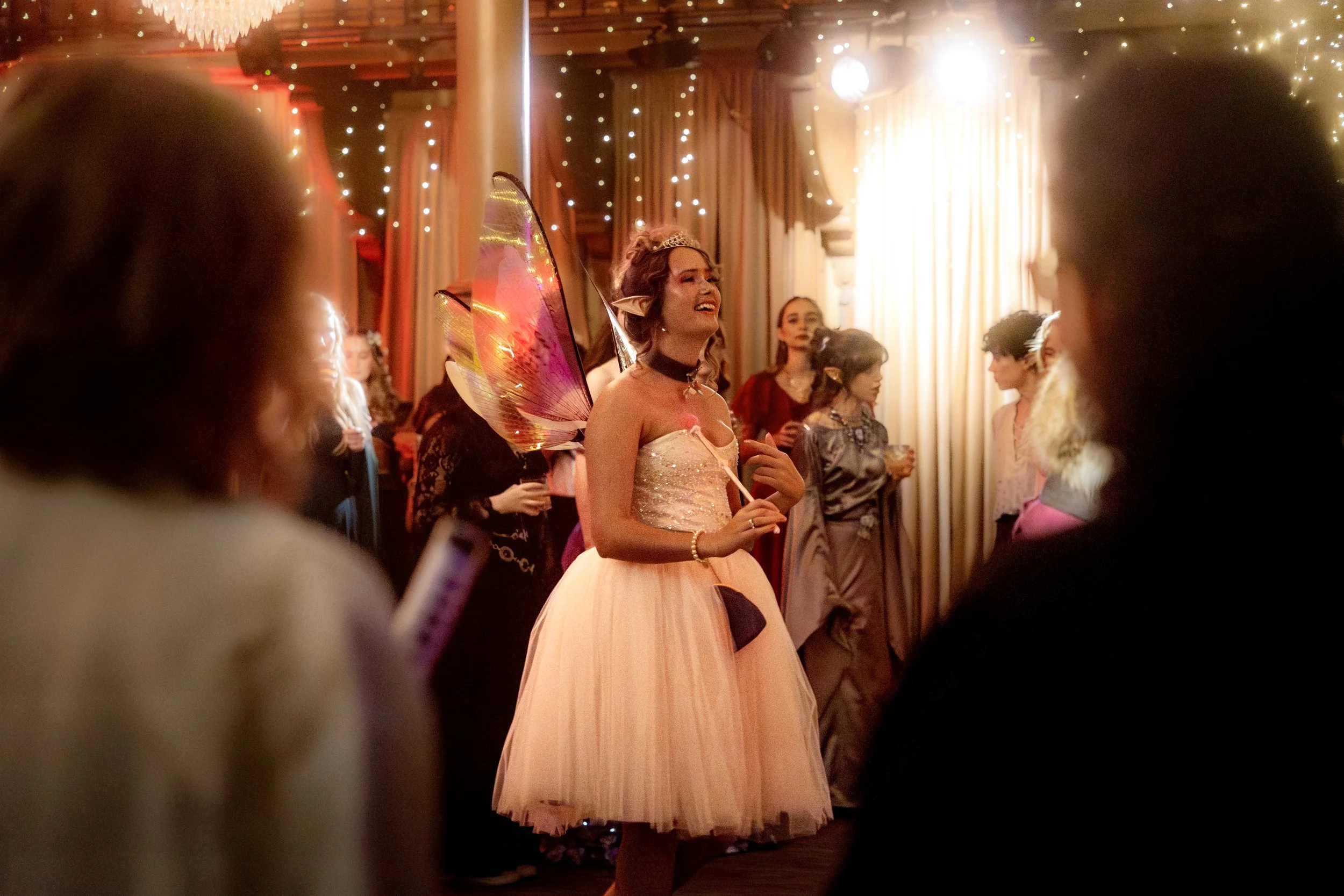 A woman dressed as a fairy or elf with butterfly wings, wearing a pink dress, standing in a festive, fairy-tale-like party scene surrounded by other women in elegant attire and fairy costumes, with warm lighting and twinkling string lights.