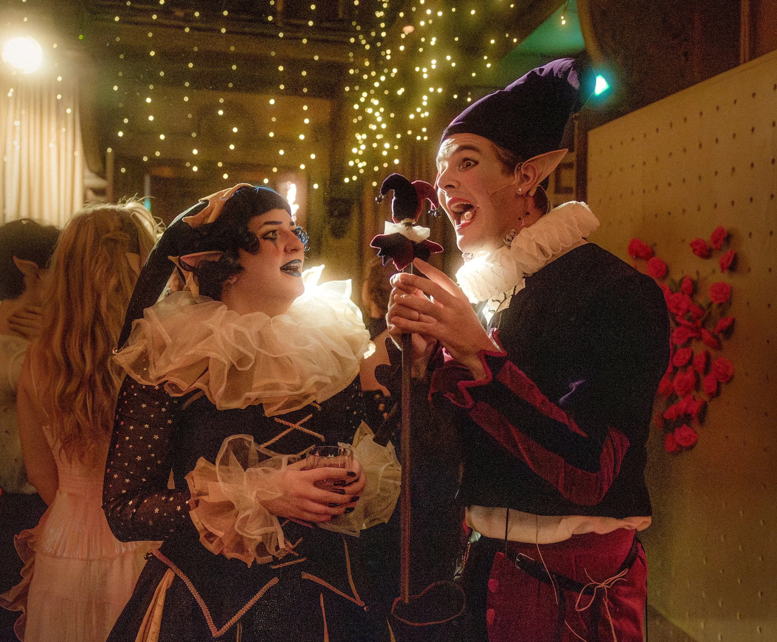 Two people dressed as elves or fairies with pointed ears and elaborate costumes are talking at a party or celebration, with one holding a drink and the other holding a flower stick. There are fairy lights and other guests in the background.