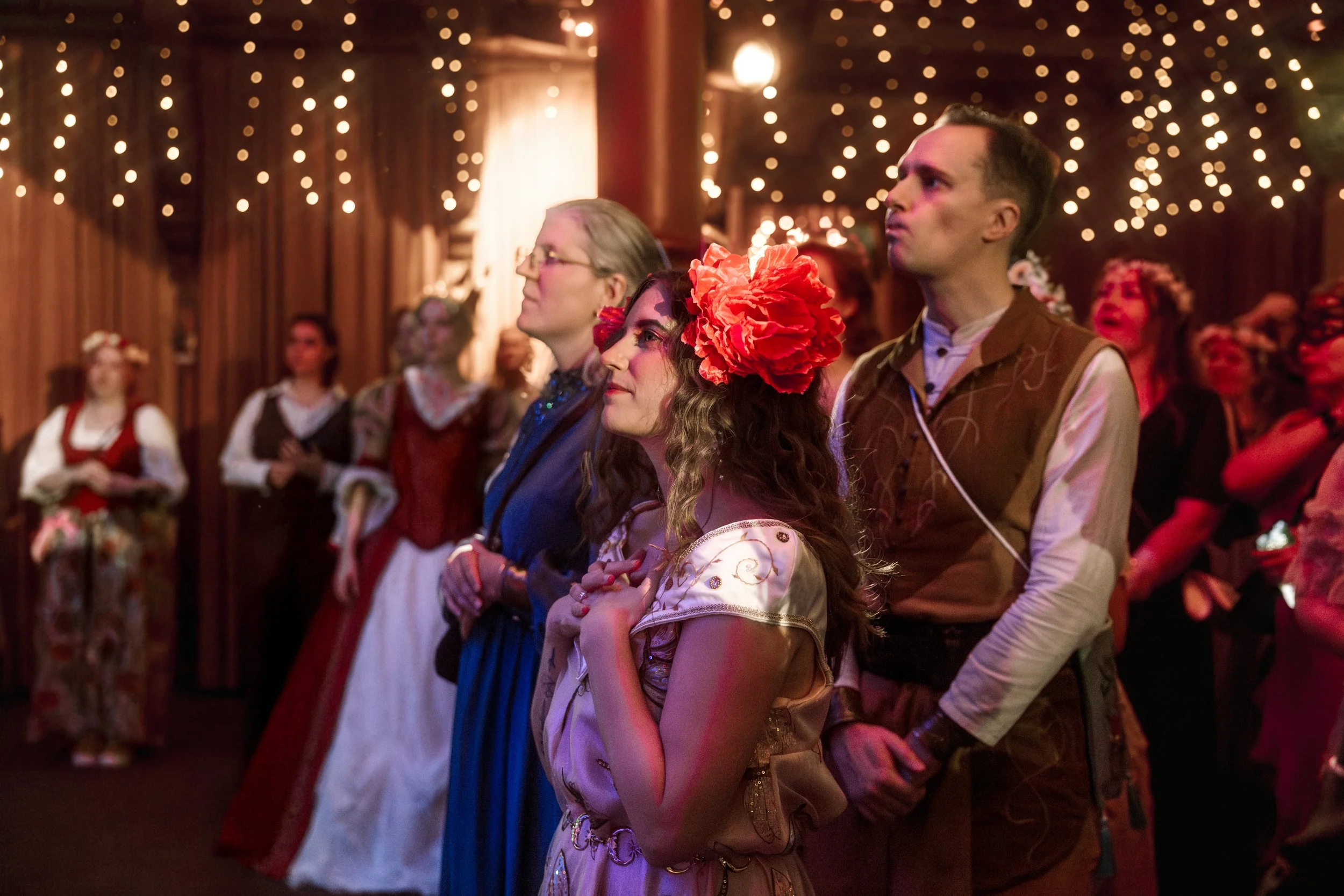 People attending an event in traditional clothing, with string lights hanging from the ceiling, in a cozy indoor setting.