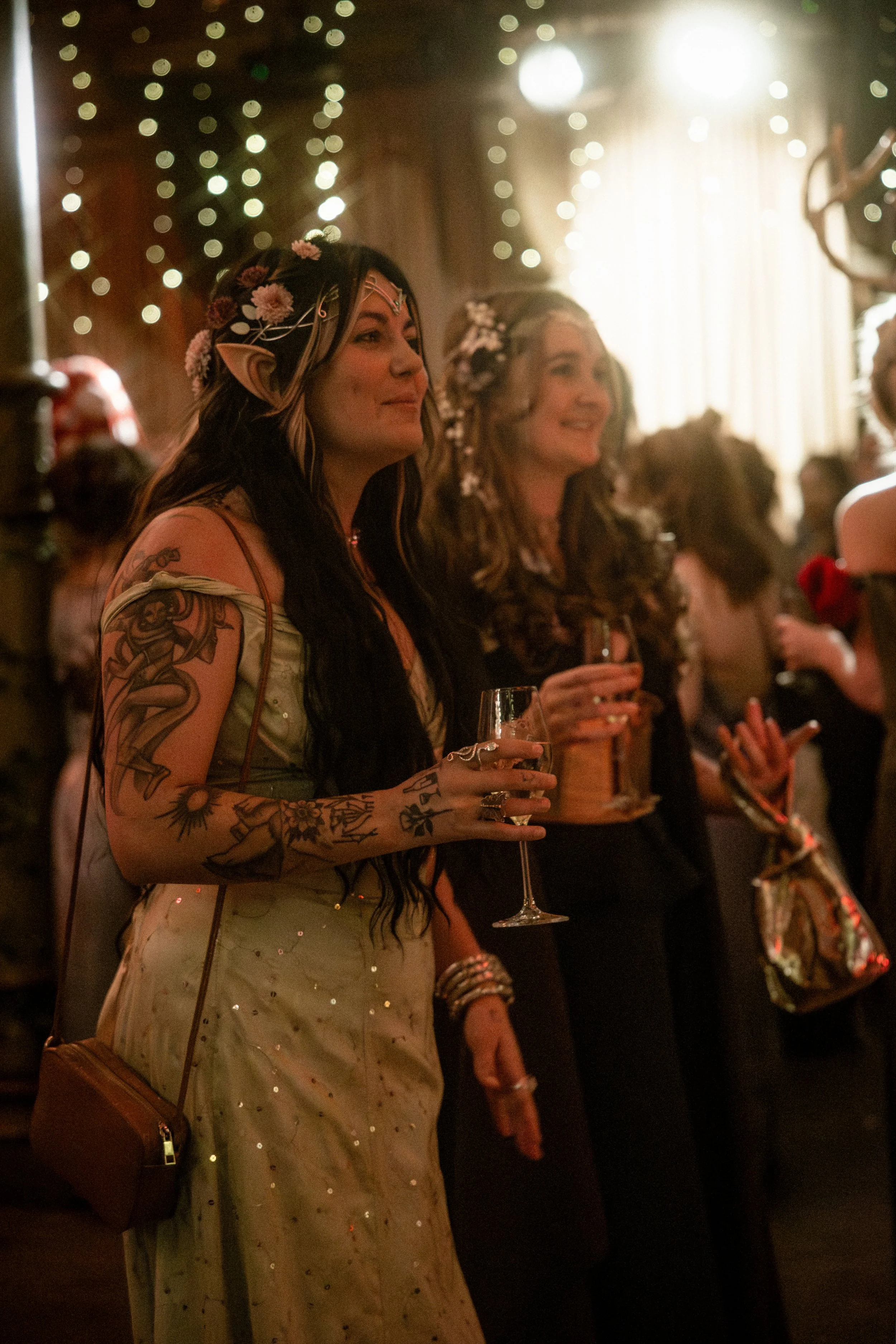 In a room lit by fairly lights, two women dressed in fantasy ball gowns are talking