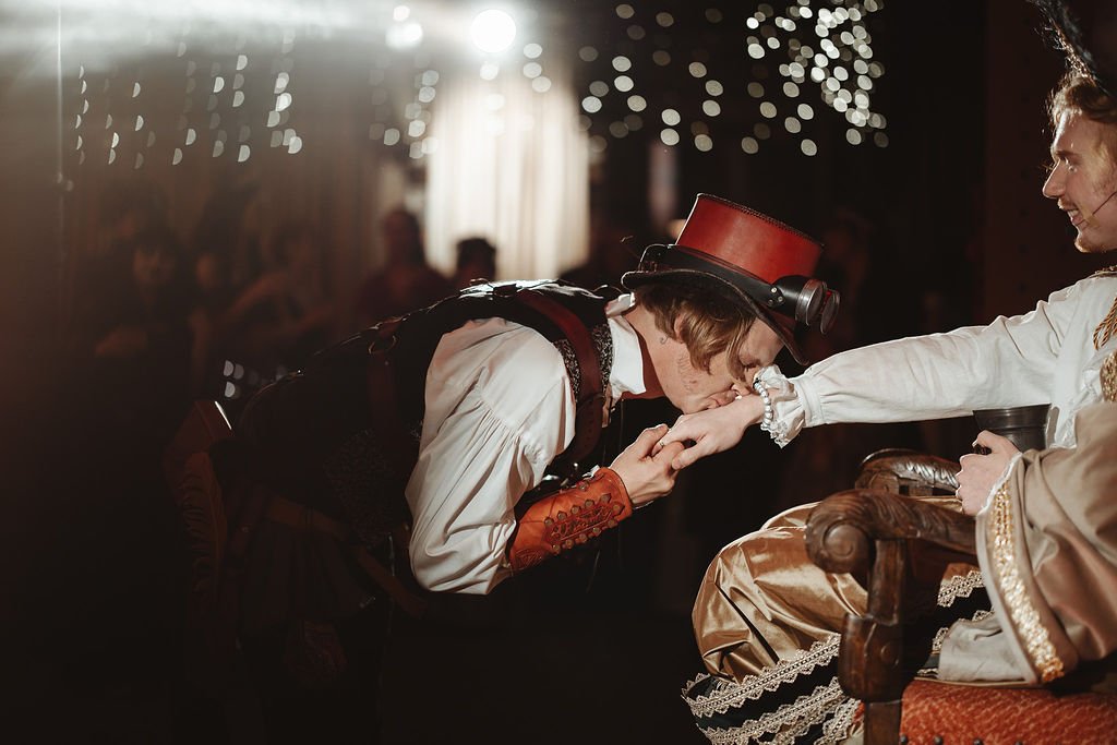 A person dressed in steampunk attire kisses the hand of another person dressed in historical costume, in what appears to be a theatrical or festive setting with blurry lights and other people in the background.