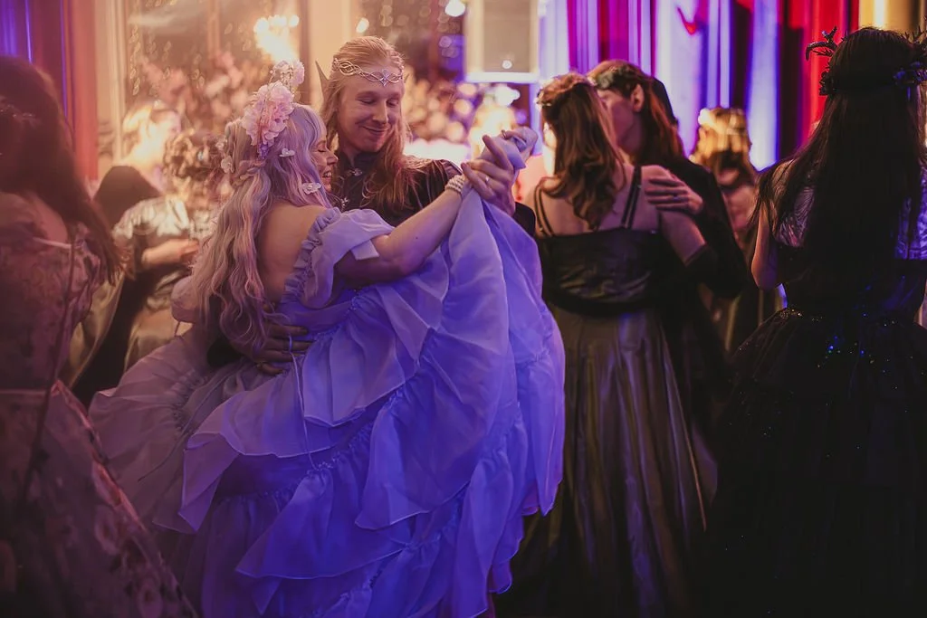 A romantic shot of a couple dancing a waltz, both dressed in ball attire, surrounded by other fantasy creatures.