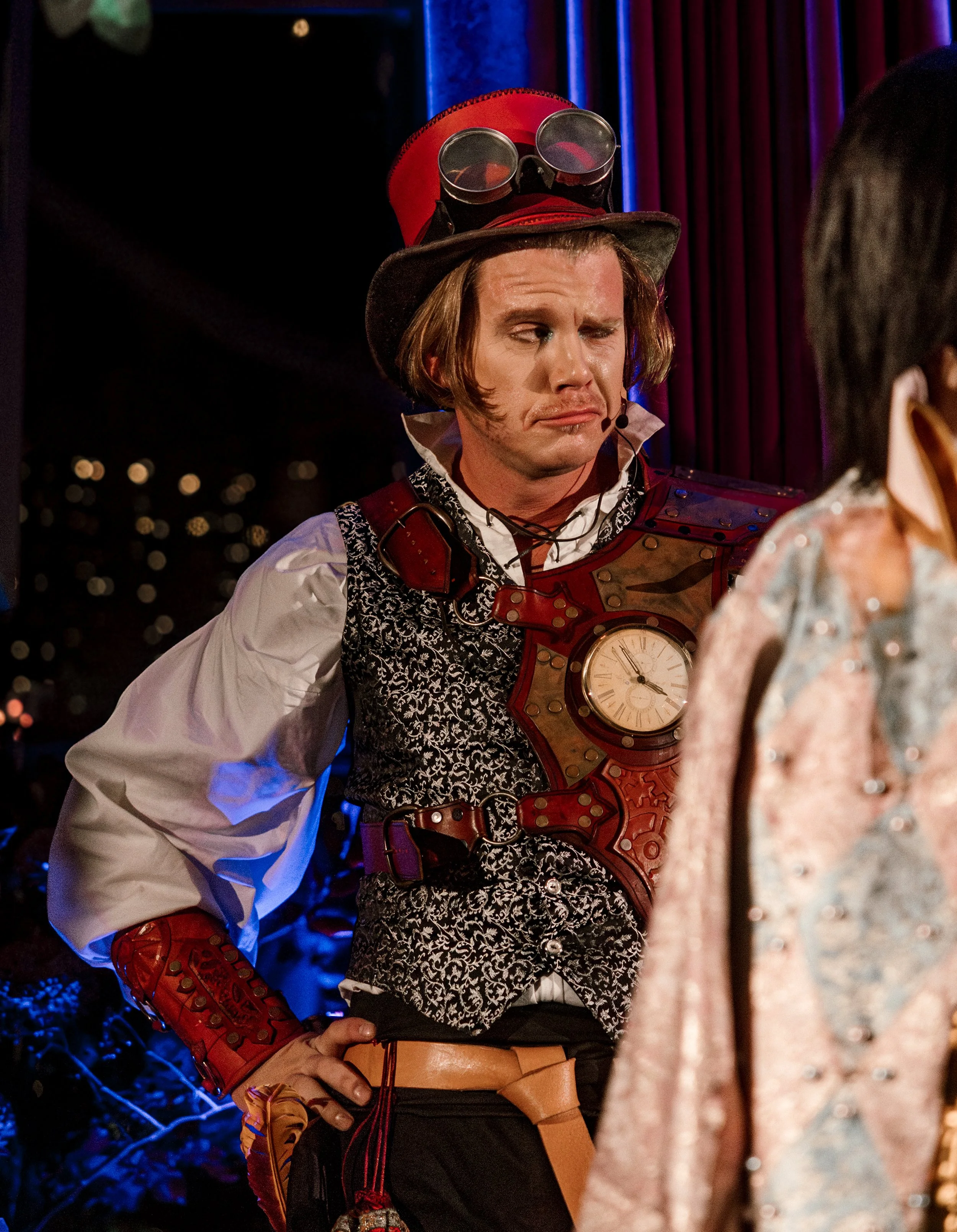 A steampunk man with leather clockwork, a tophat with goggles and smart attire talks to a guest at the ball.