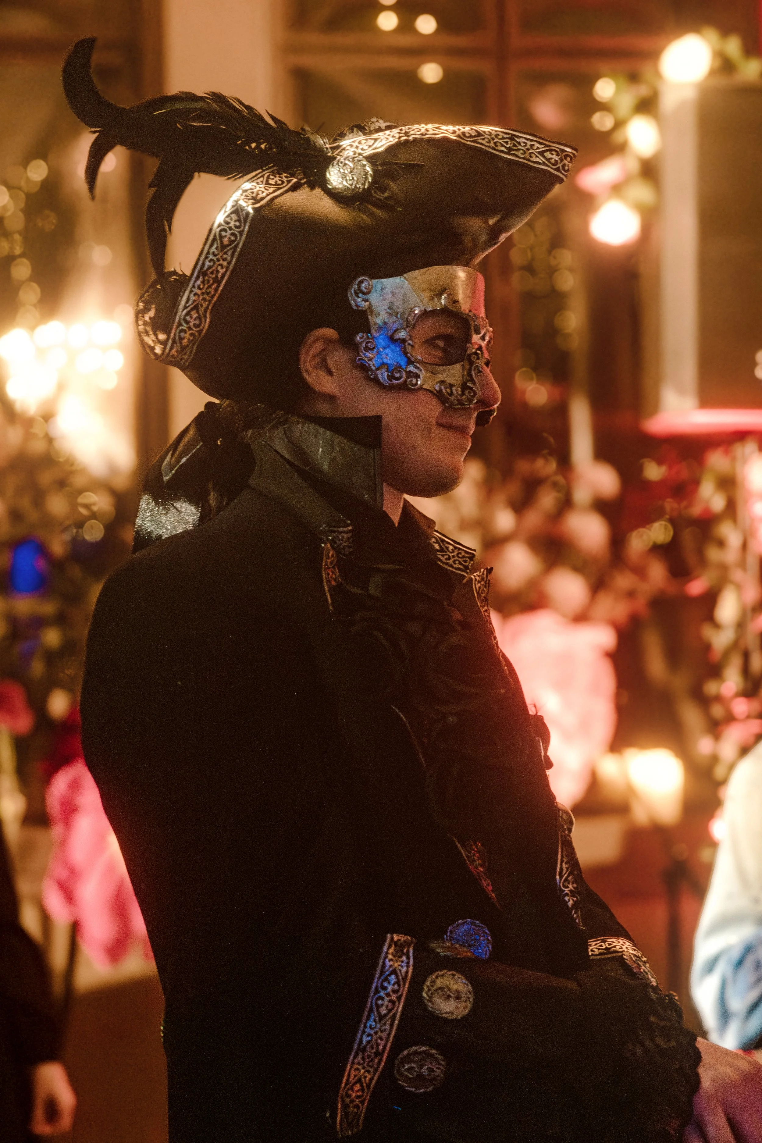 Person dressed in a colorful, ornate costume with a large hat decorated with feathers, wearing a decorative mask, at a festive event with warm lighting and blurred background of other attendees.