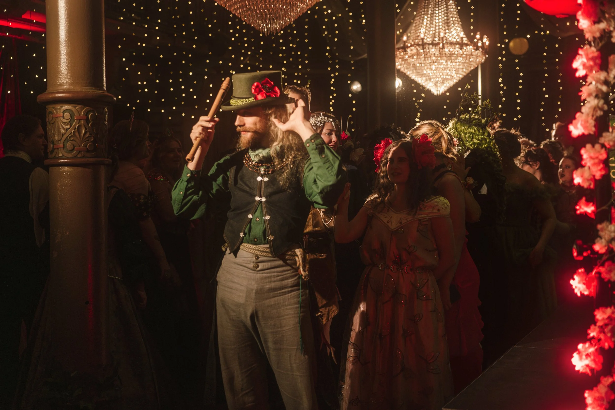 A man dressed in a vintage costume wearing a hat, surrounded by women in colorful costumes and floral headpieces, at a festive event with warm lighting, chandeliers, and decorative lights.