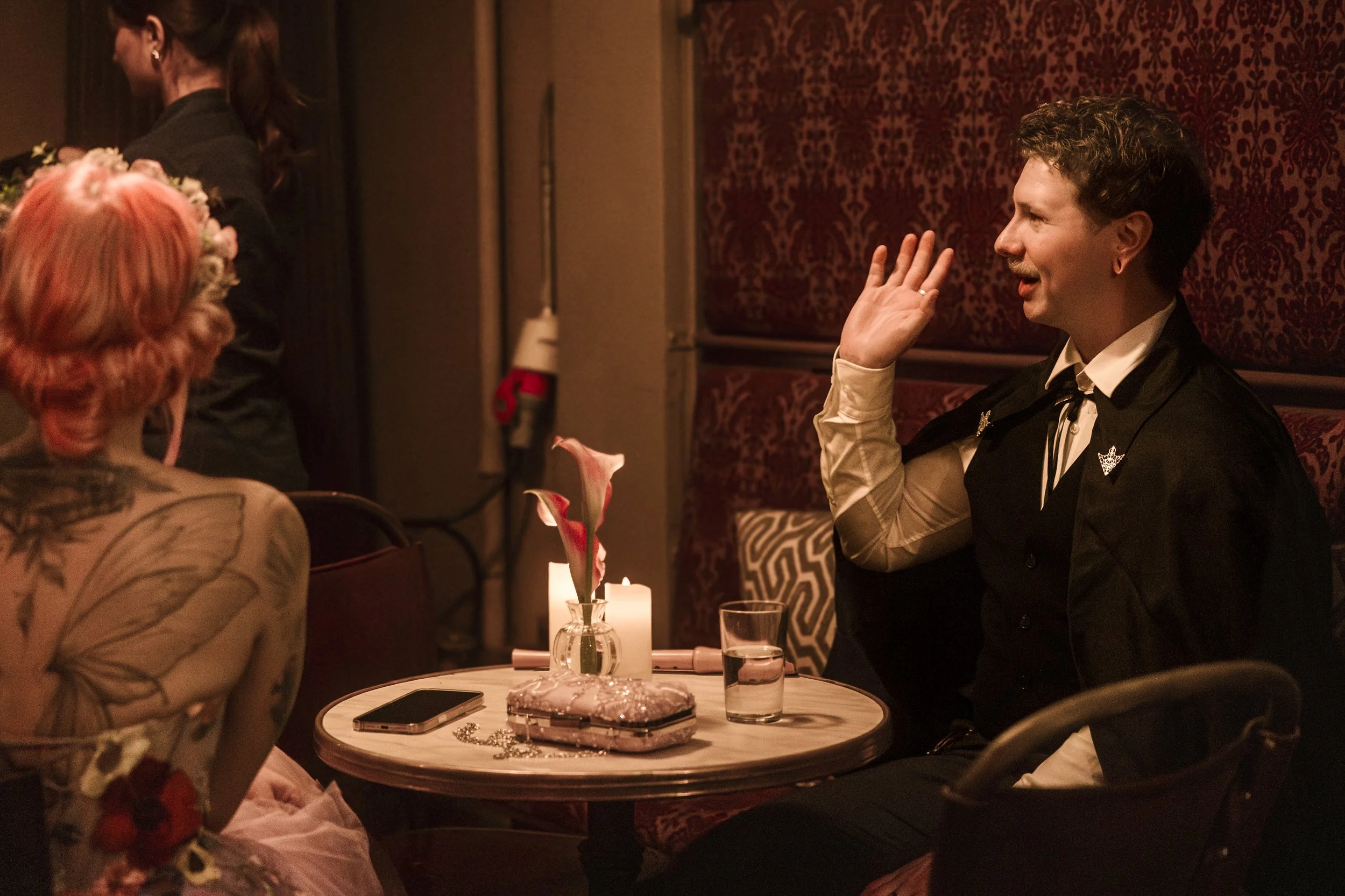 A man with short curly hair and a mustache wearing a dark vest and a white shirt is sitting at a round table, smiling and waving. In front of him, a woman with pink hair and tattoos on her shoulder is sitting, facing away. The table has a vase with p