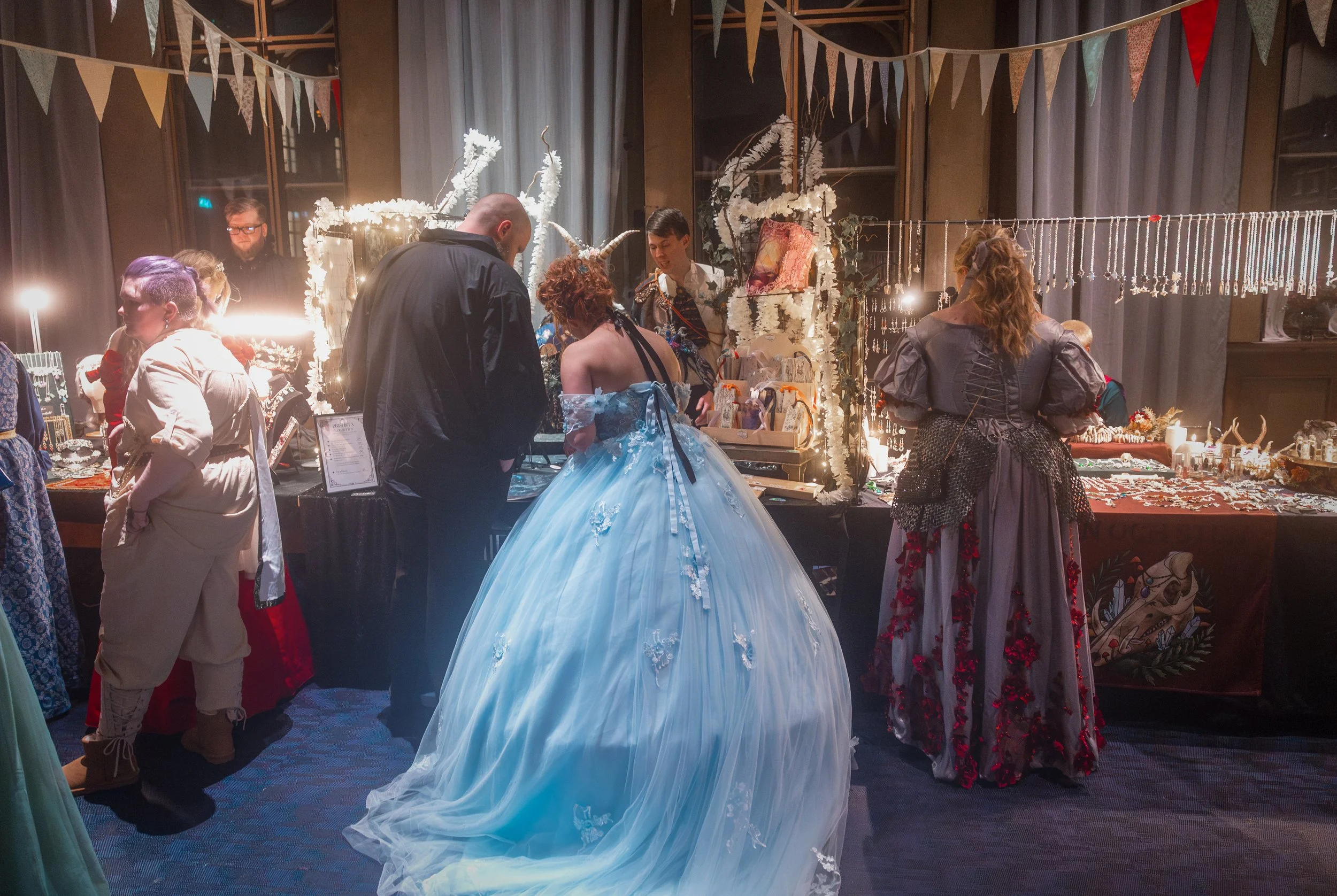 People browsing jewelry and art pieces at an indoor market stall decorated with fairy lights and bunting.