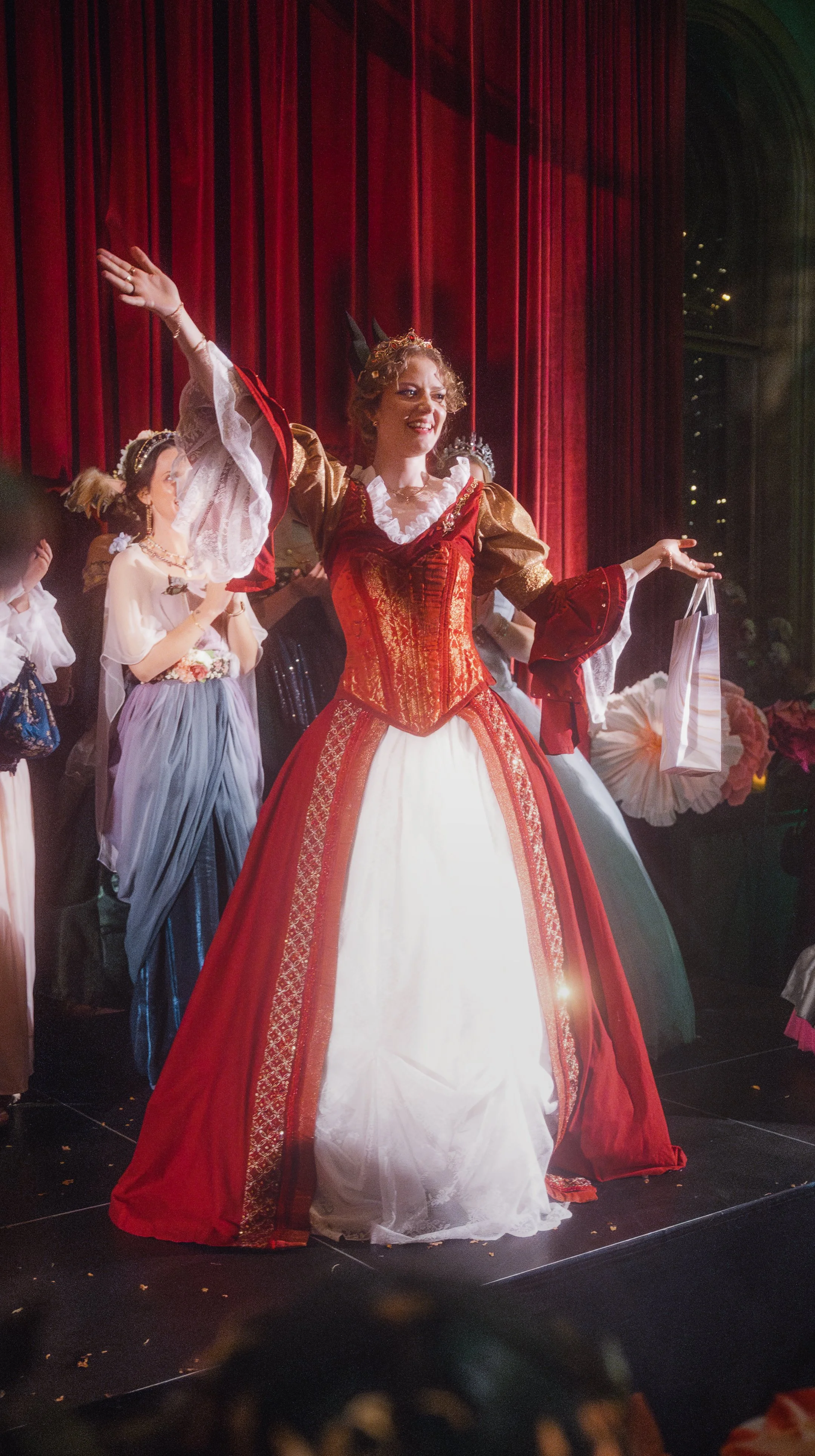 Woman in a red and white costume with a corset and a crown on her head, standing on stage with others in costumes, under red curtains.