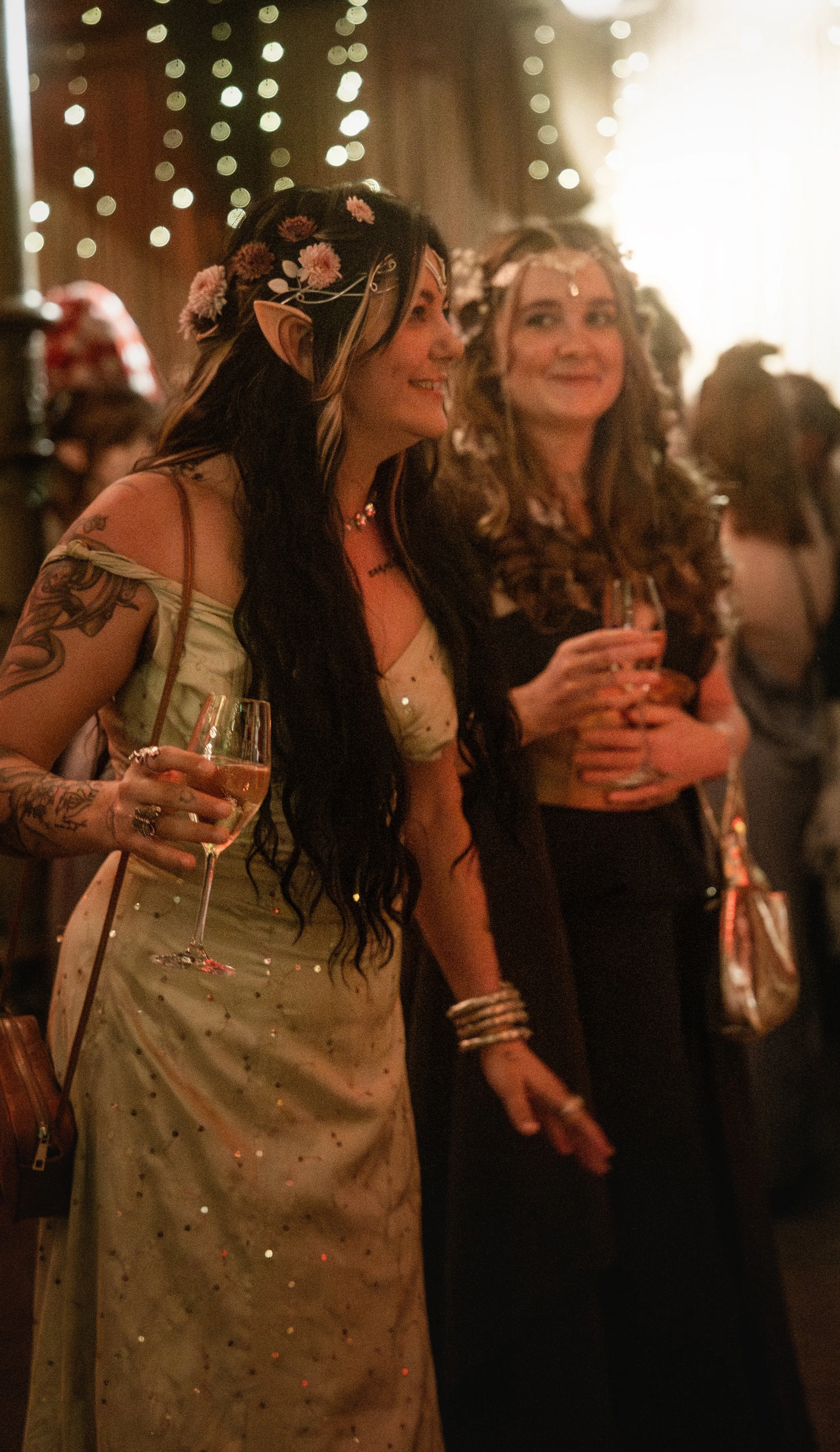 In a room lit by fairly lights, two women dressed in fantasy ball gowns are talking