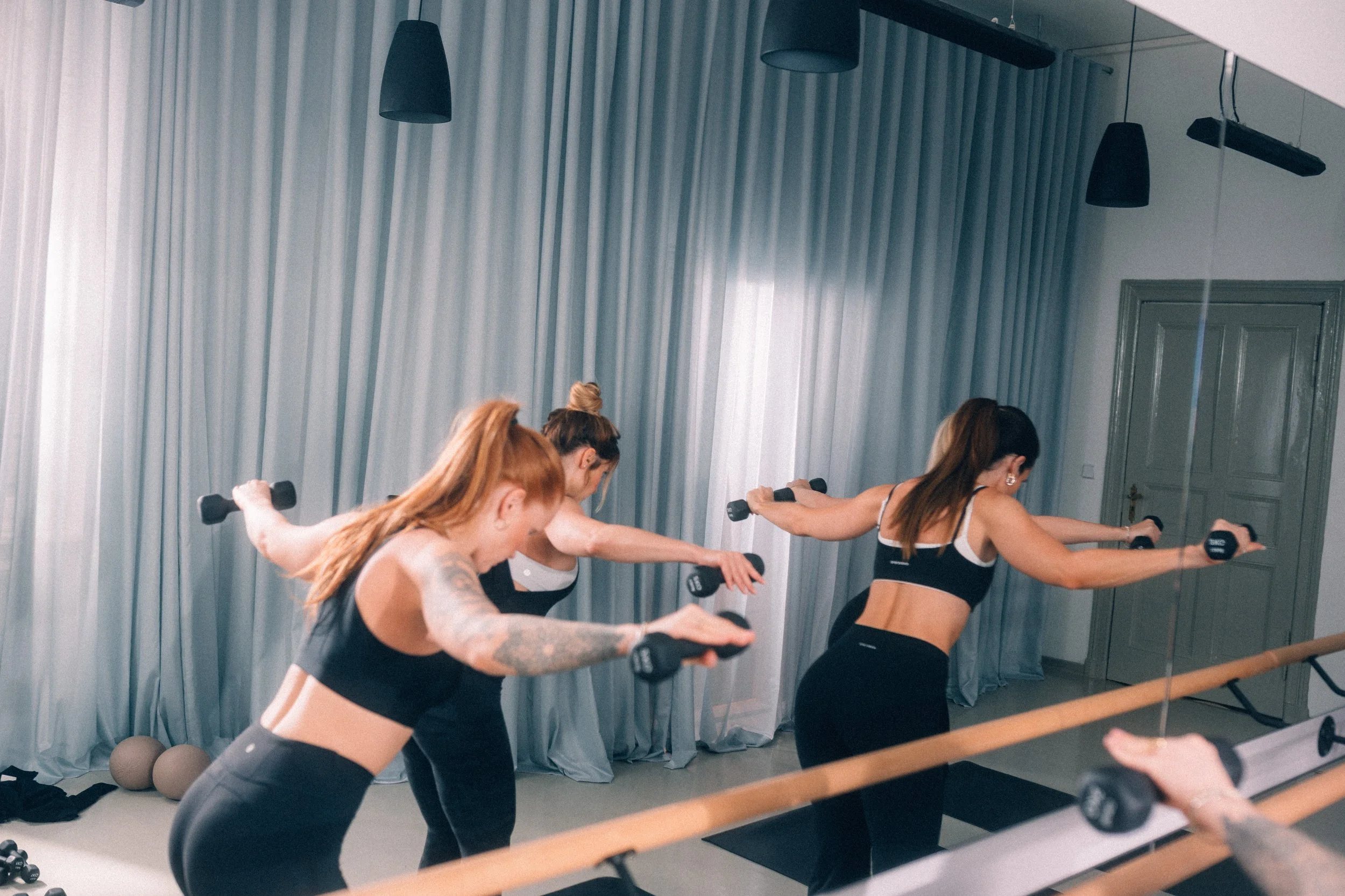 Mehrere Frauen beim Workout im Fitnessstudio, die Hanteln heben, vor Spiegel, in Sportkleidung.