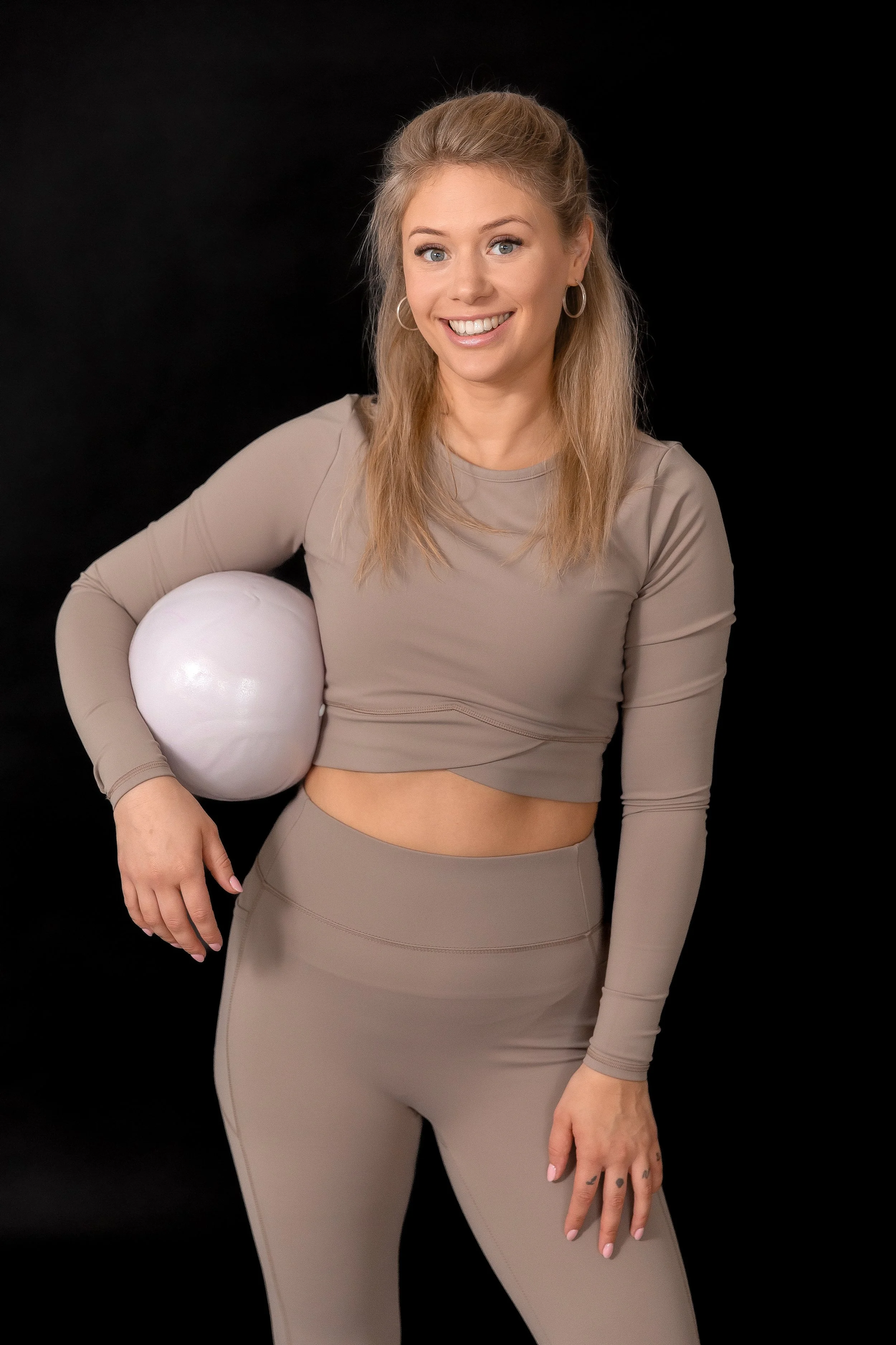 Eine lächelnde Frau trägt einen beige Sport-BH und Leggings, hält einen weißen Ball unter dem Arm, vor schwarzem Hintergrund.
