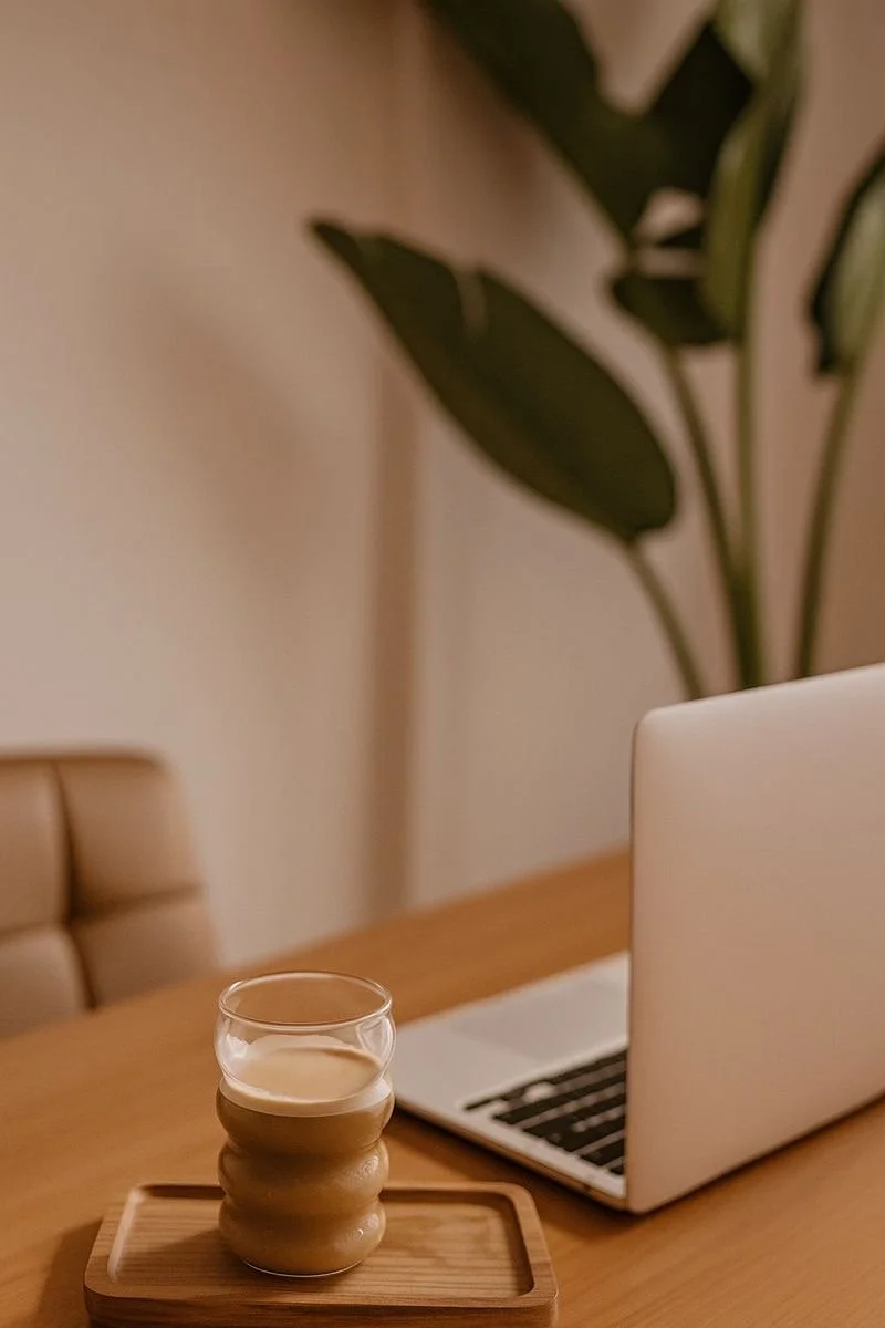 Ein Tisch mit einem Laptop und einem Glas Milch auf einem Holztablett, im Hintergrund eine große Pflanze in einem Raum mit beige Wänden.