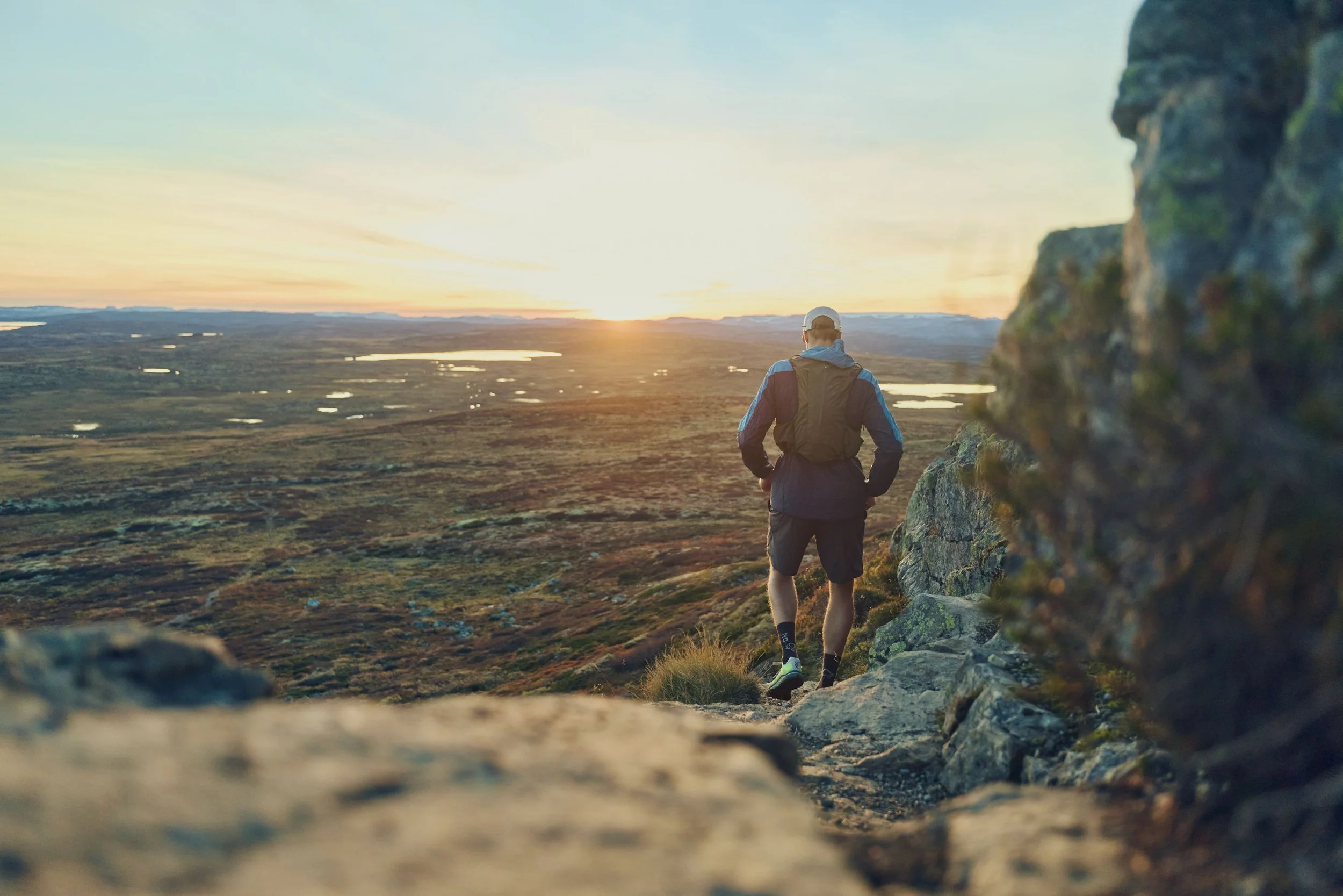 A man hiking on a trail with a backpack, walking towards a sunset over a vast, wetland landscape with small lakes and greenery.