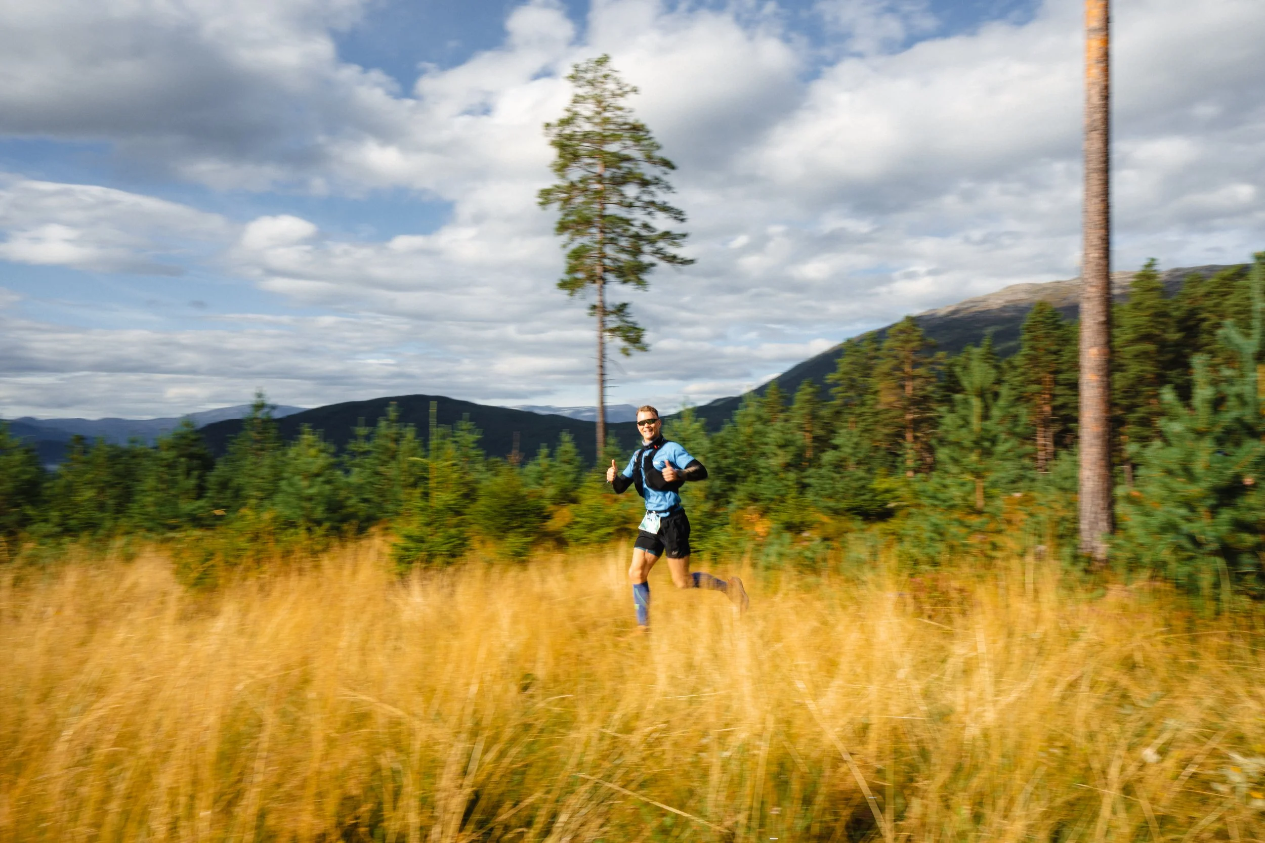 A man trail running through tall grass in a scenic outdoor landscape with trees, mountains, and a partly cloudy sky.