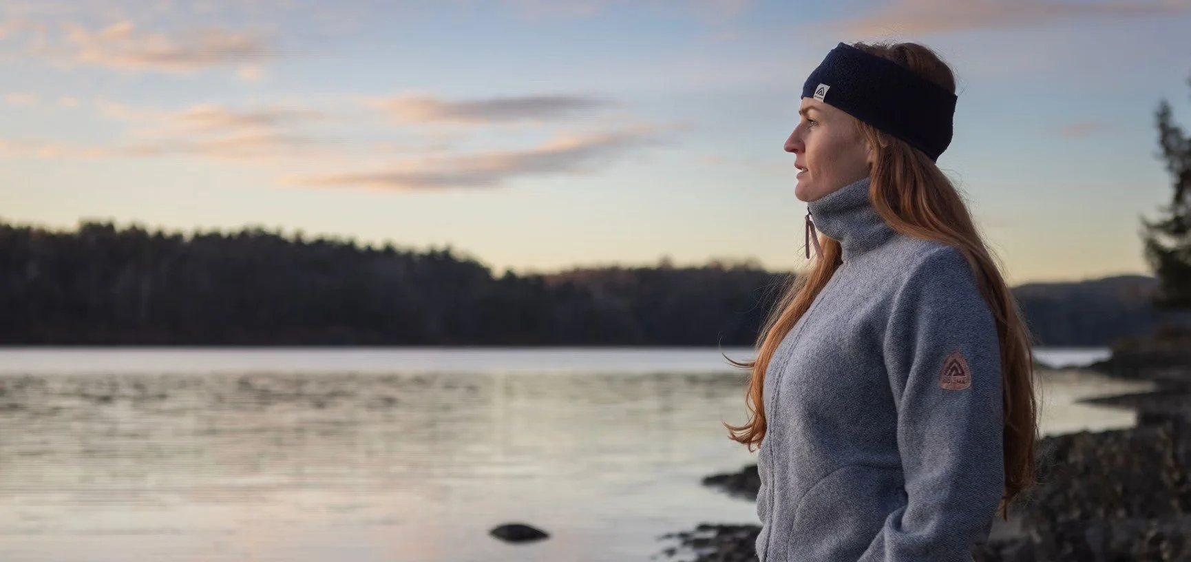 A woman with long red hair standing near a body of water during sunset, wearing a gray outdoor jacket and a black headband.