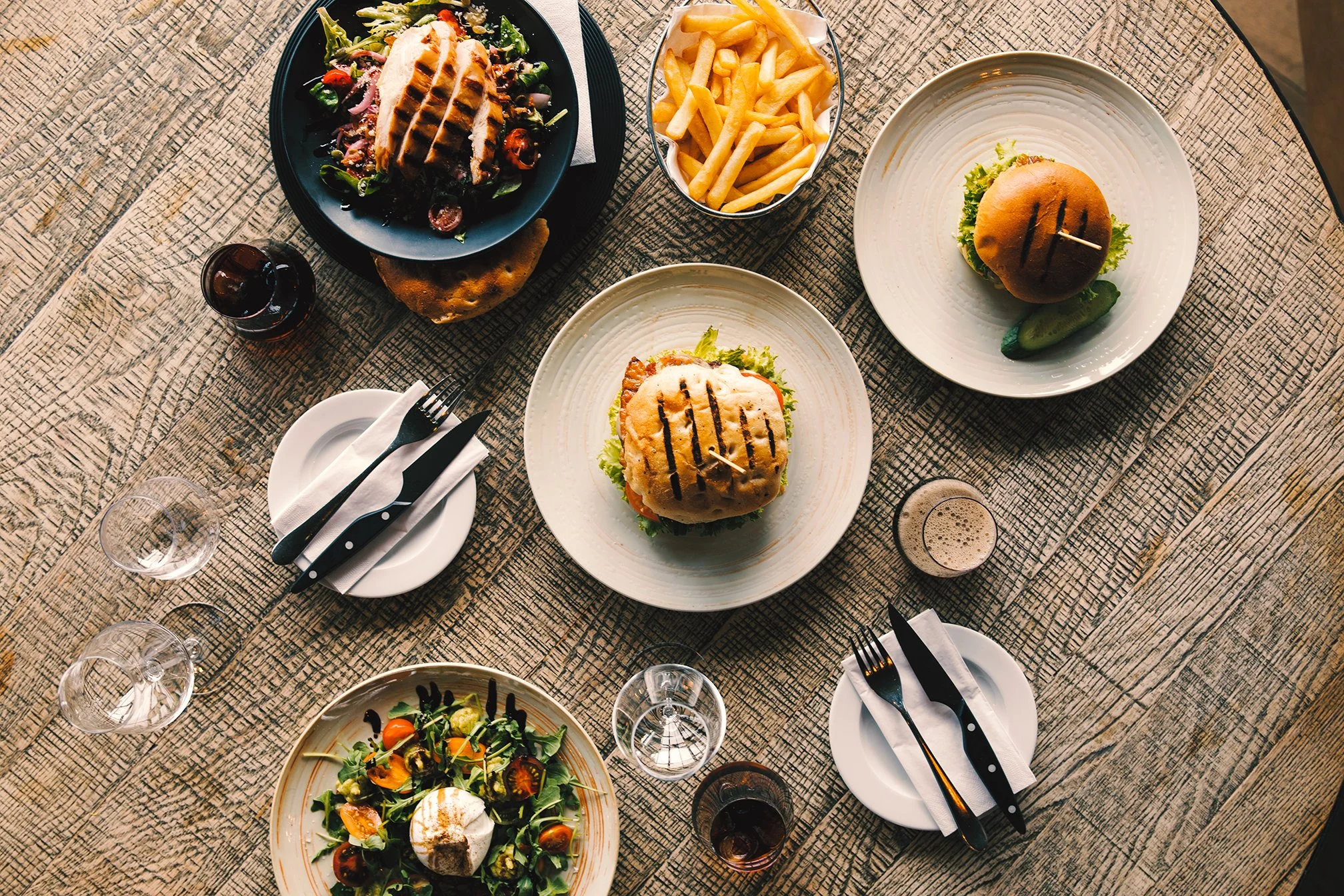 A top-down view of a table set with various dishes and drinks, including salads, sliders, fries, a burger, a sandwich, and glasses of water and soda on a rustic wooden table.