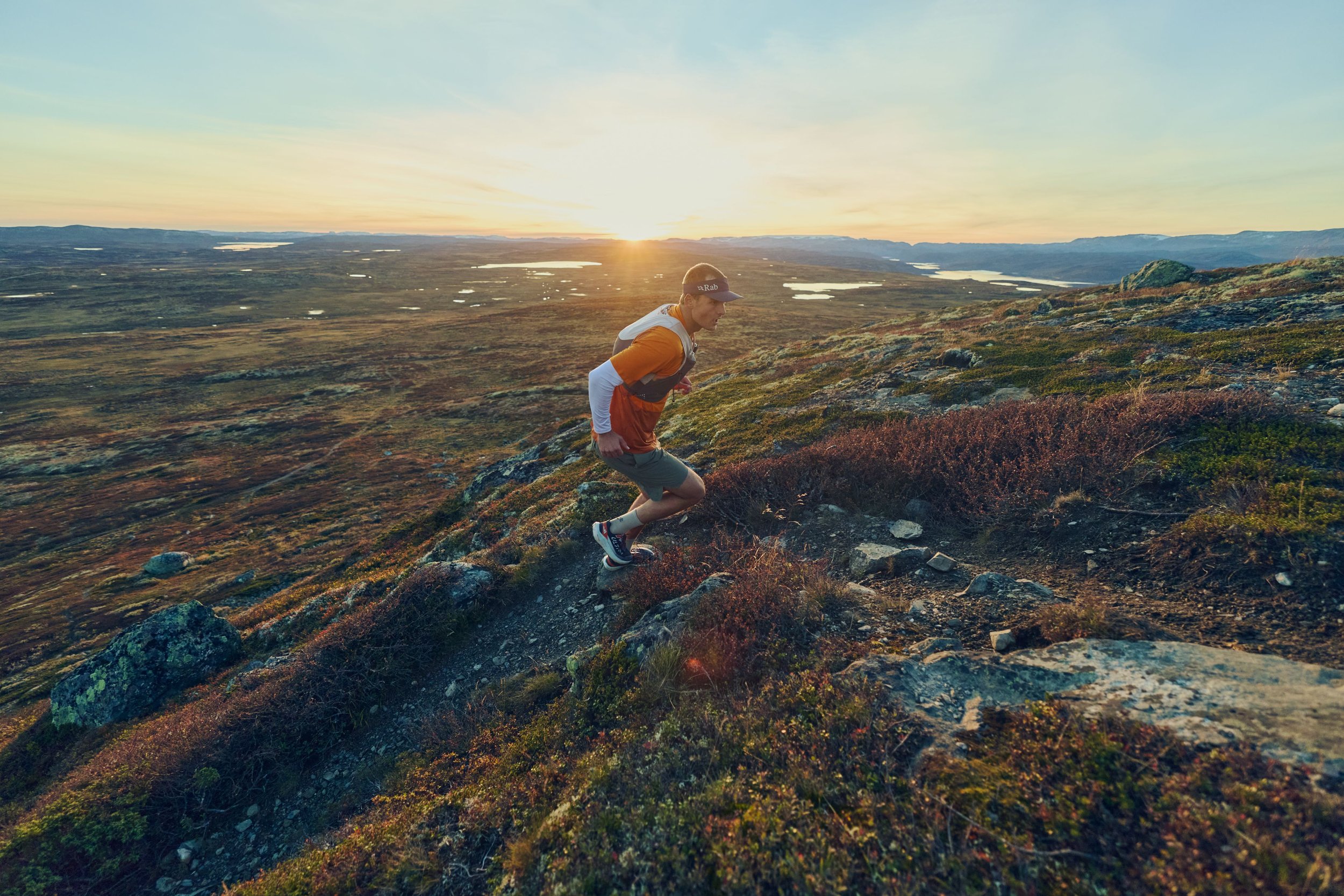 Person trail running on a rugged mountain path at sunset, with a wide landscape of lakes and hills in the background.