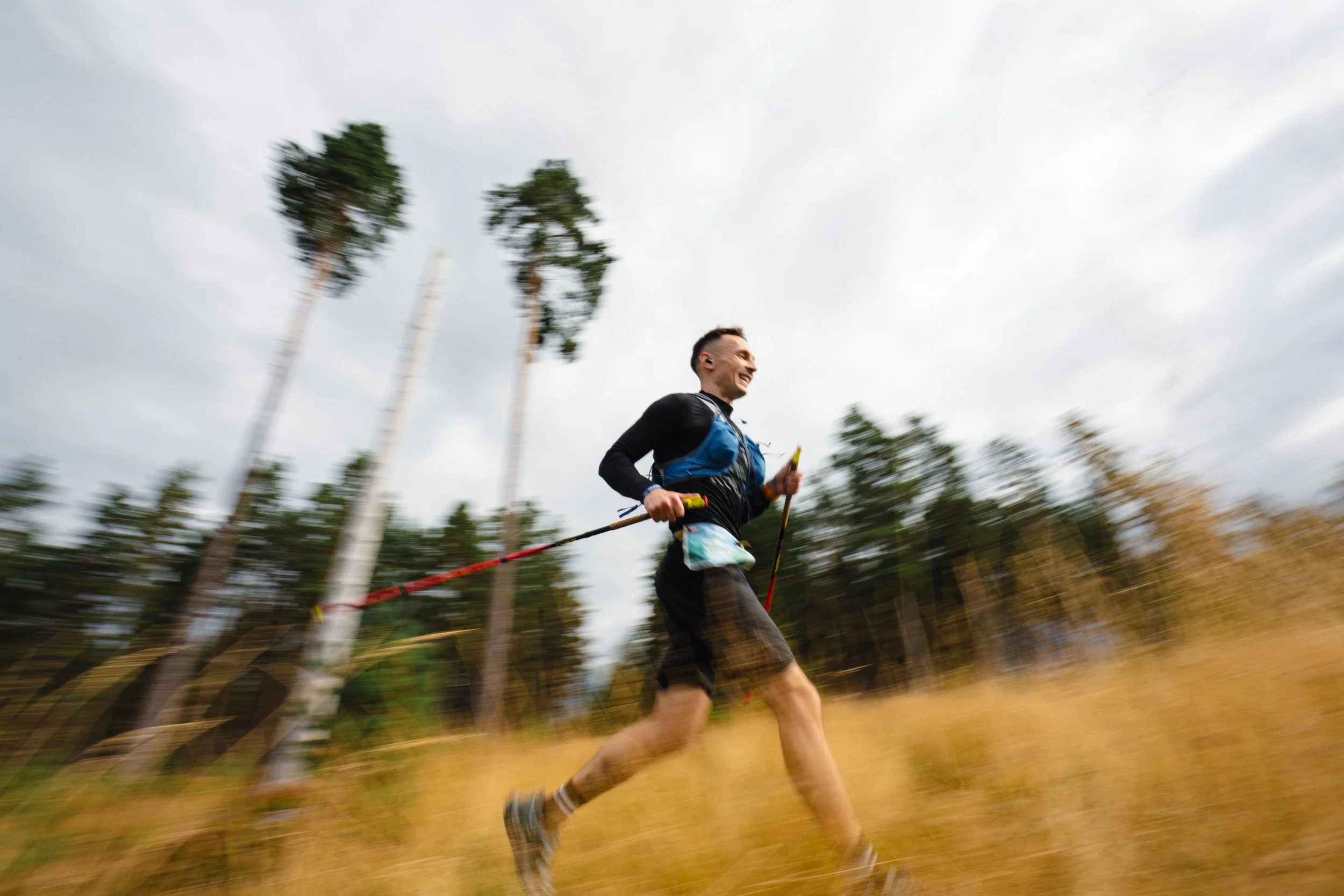 A man trail running through a grassy field with tall trees in the background, during overcast weather.