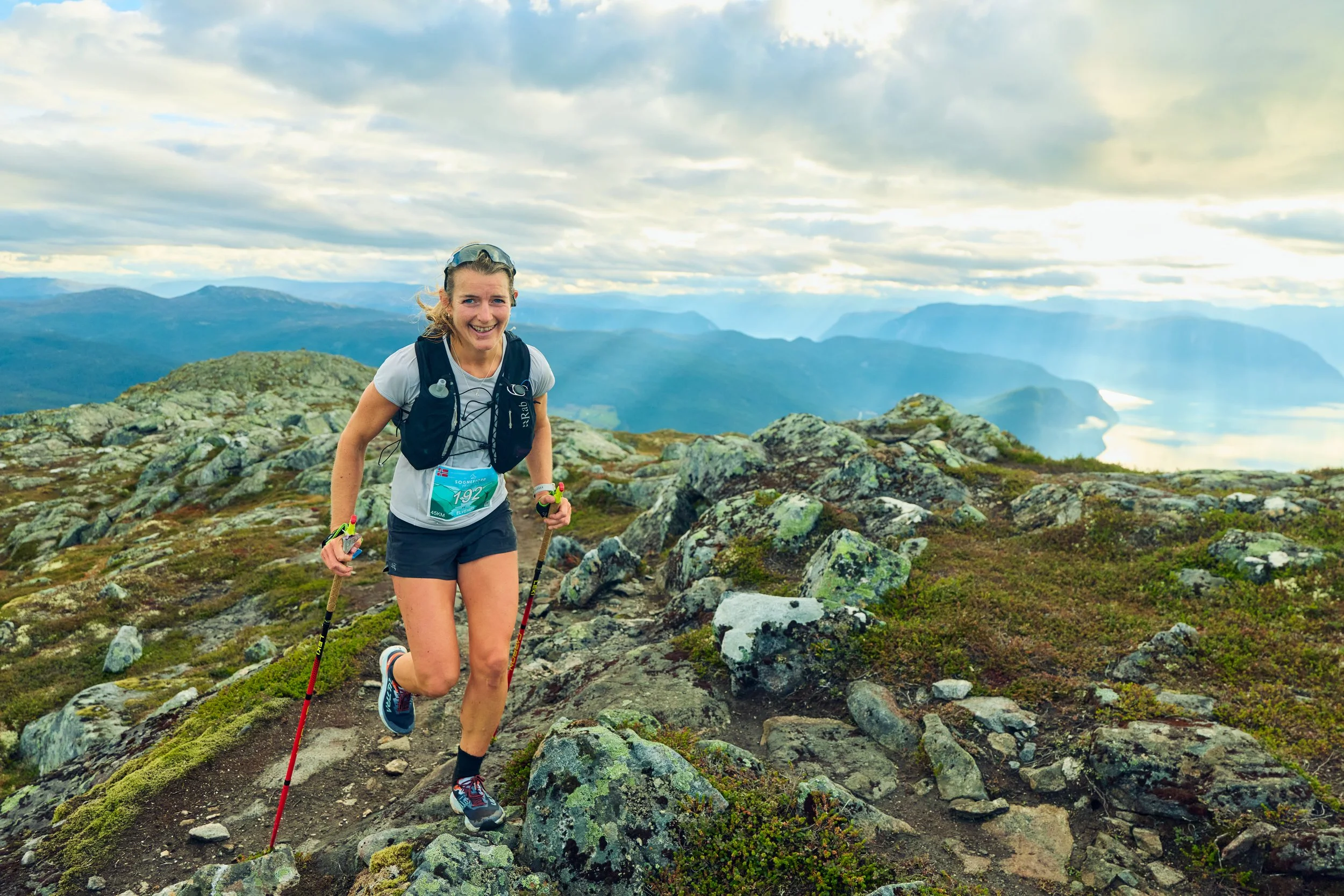 A woman trail running on a rocky mountain trail with scenic mountains and a body of water in the background, under a partly cloudy sky.