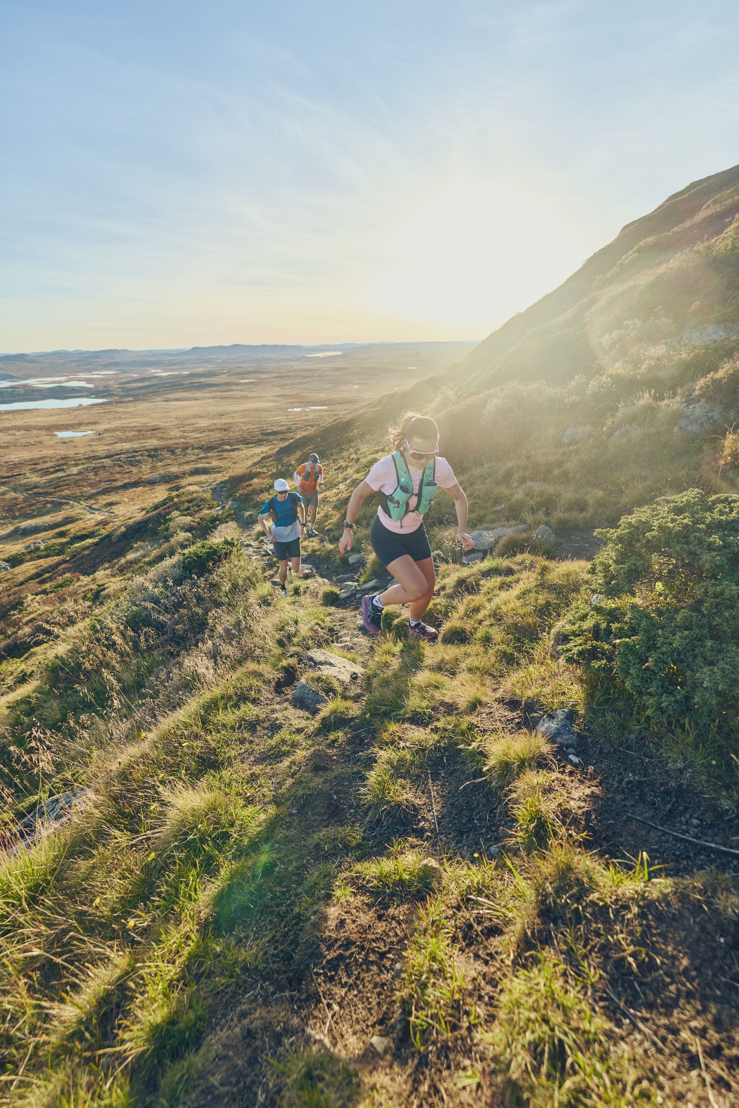 Group of hikers climbing a rugged trail on a hillside at sunset, with expansive plains and water bodies in the distance.