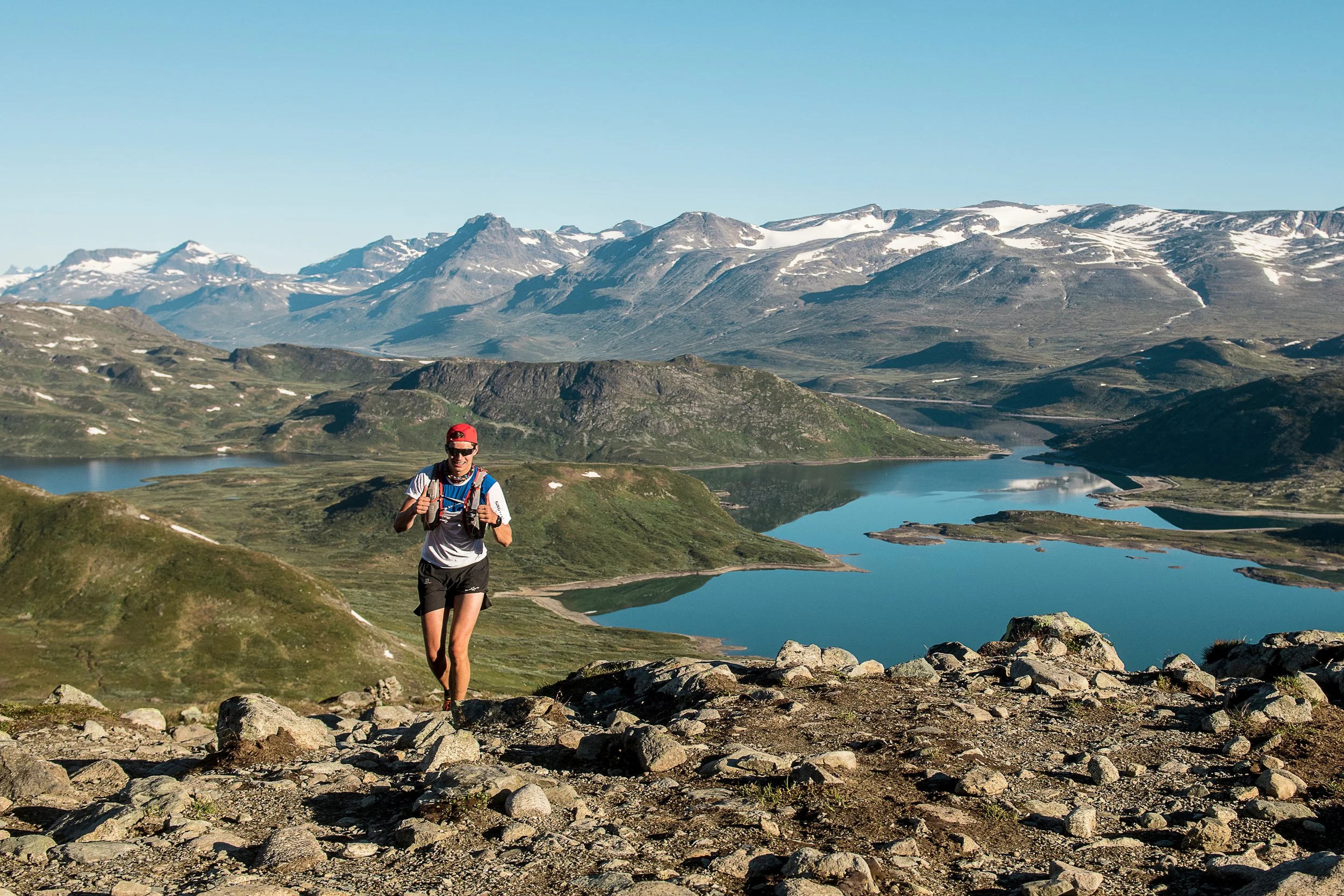 A person running on a rocky trail in a mountain landscape with lakes and snow-capped peaks in the background.