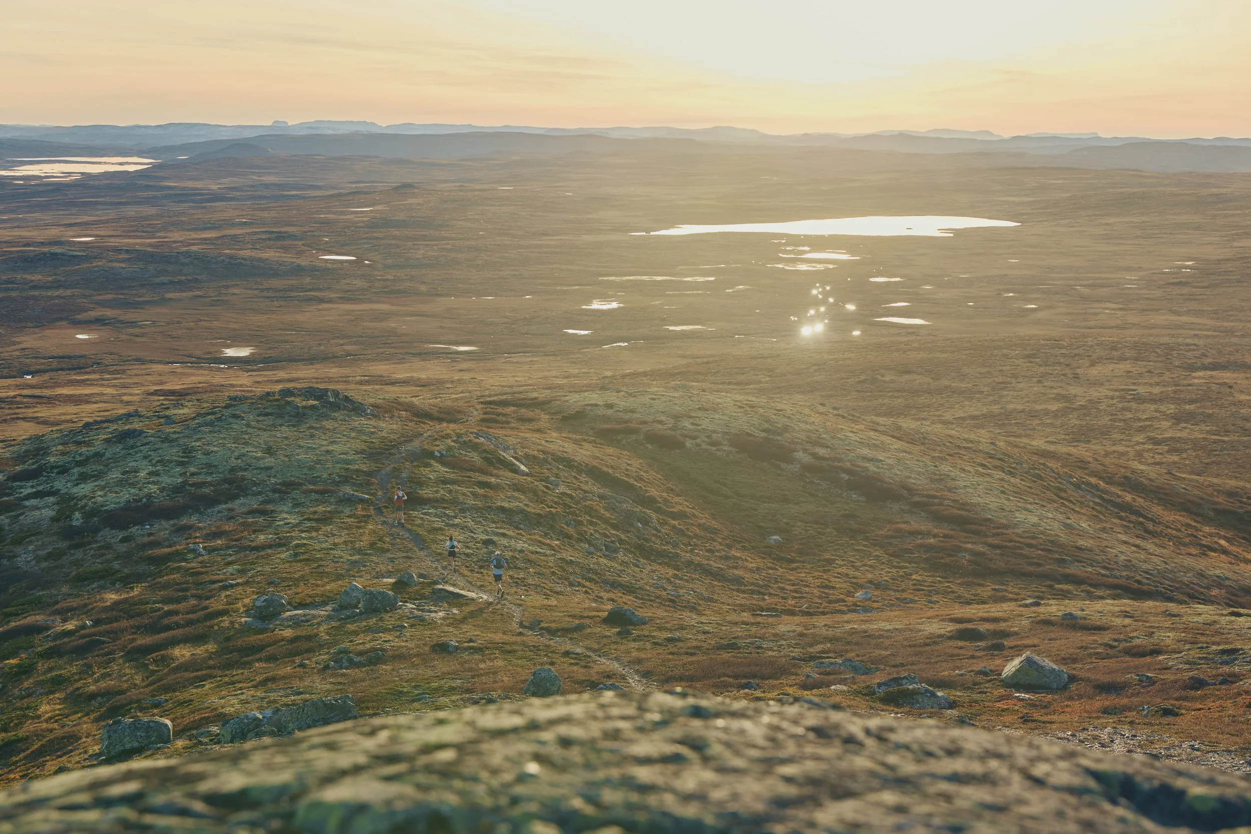 Hikers walking on a trail in a vast, open landscape at sunset, with hills, lakes, and distant mountains in the background.