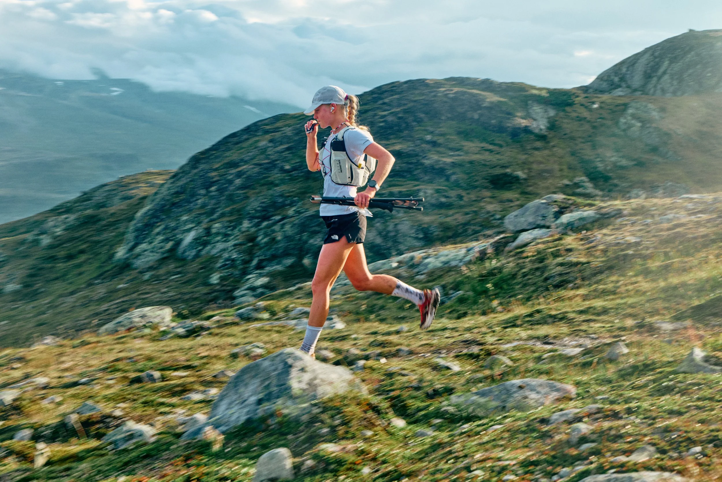 A woman trail running on a rocky mountain trail with green hills and mountains in the background, wearing a cap, shorts, and a hydration vest.