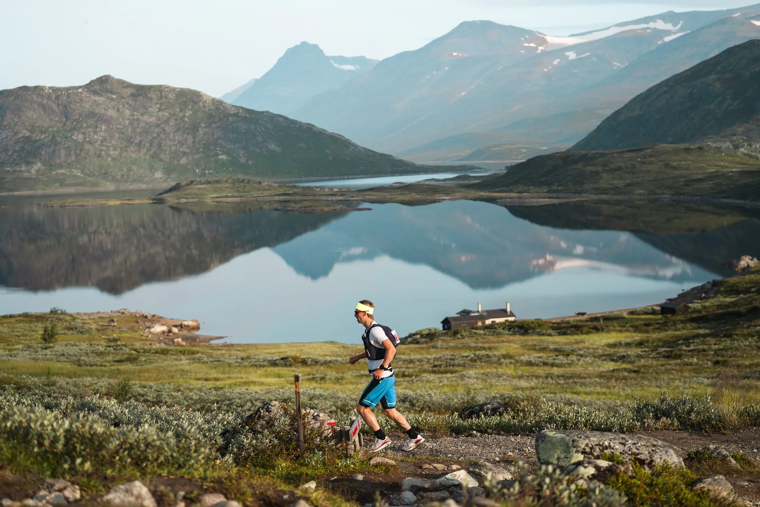 A man trail running on a rocky dirt path through a lush green landscape beside a body of water with mountains in the background.