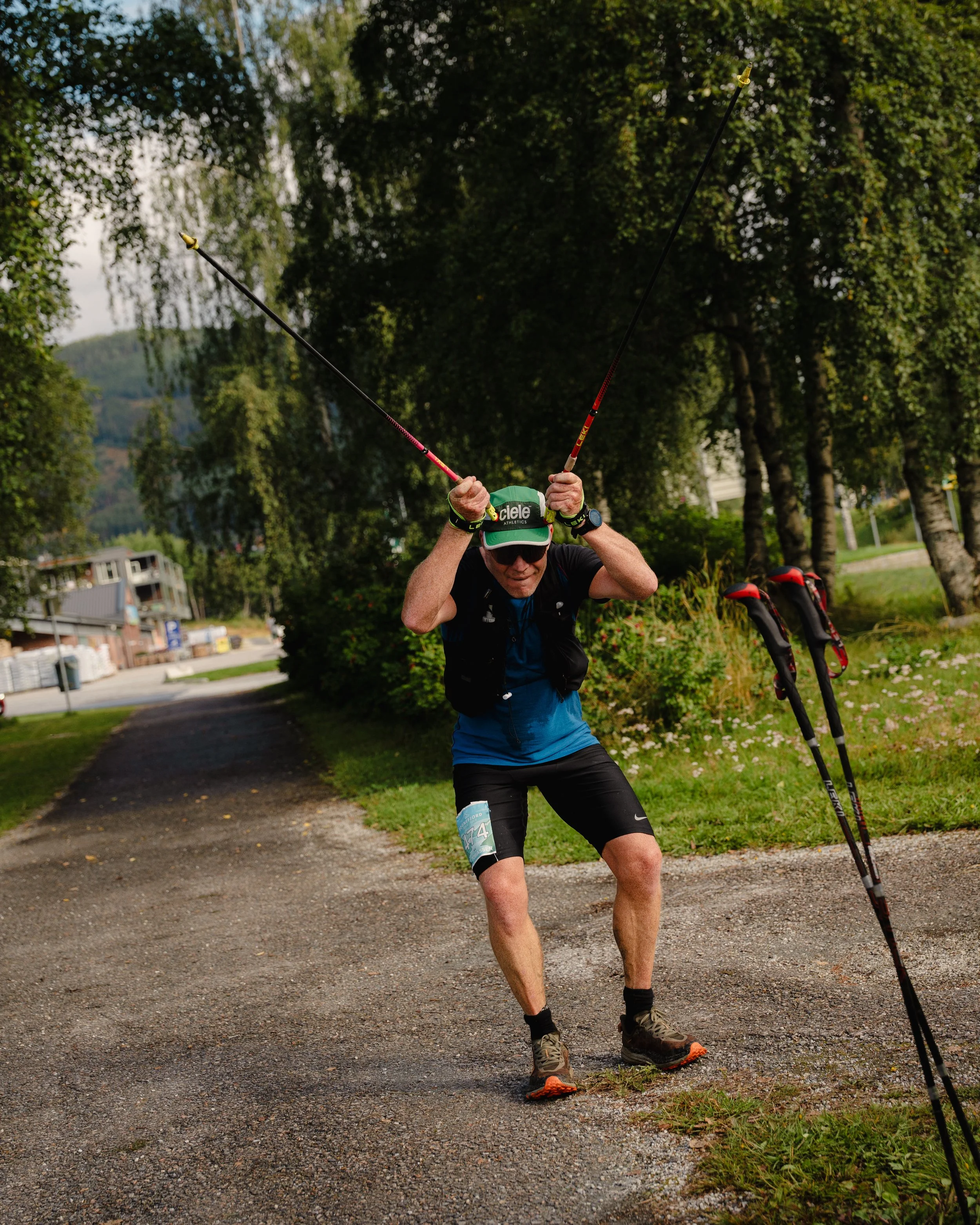 An older man in athletic gear and sunglasses celebrating during a trail run in a park, standing on a gravel trail with trees and greenery around him.