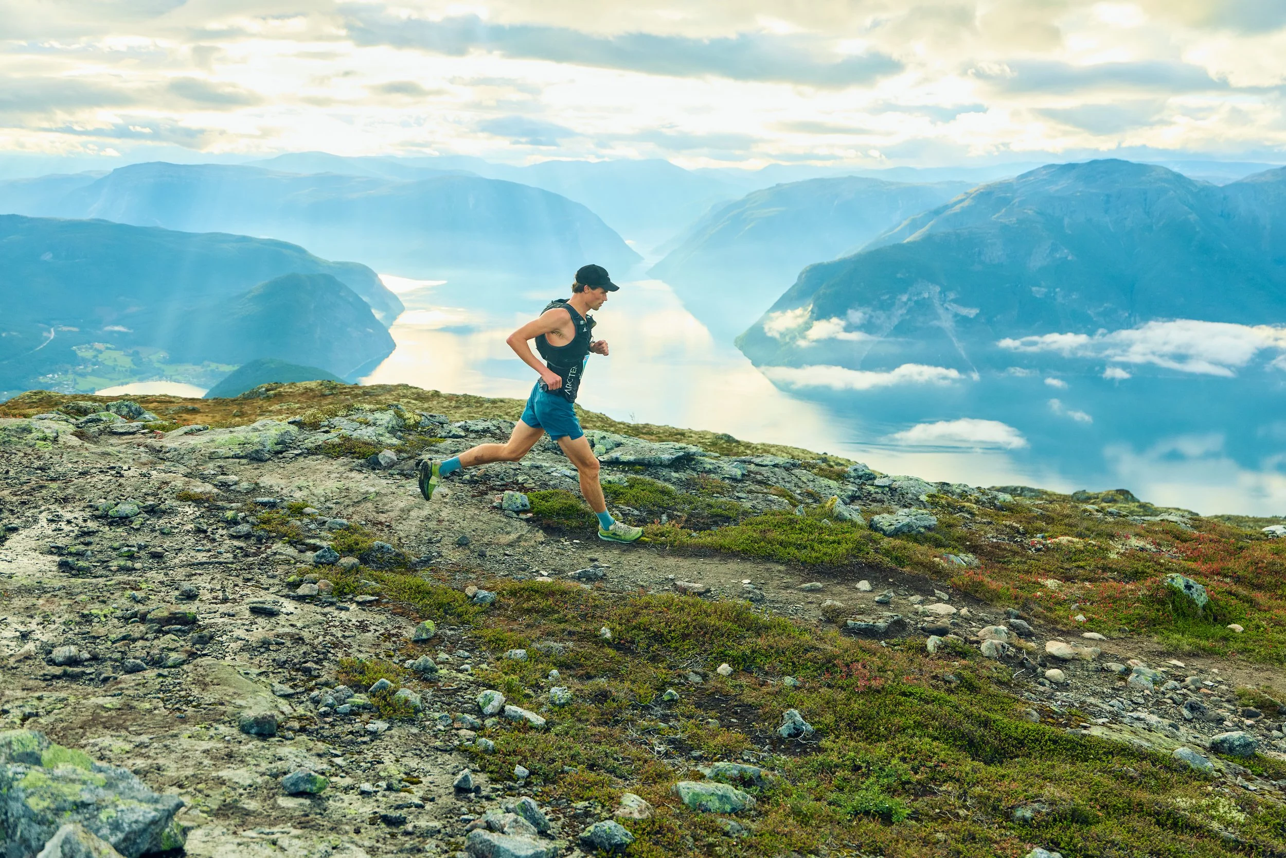 A man running on a rocky mountain trail with a scenic lake and mountains in the background.