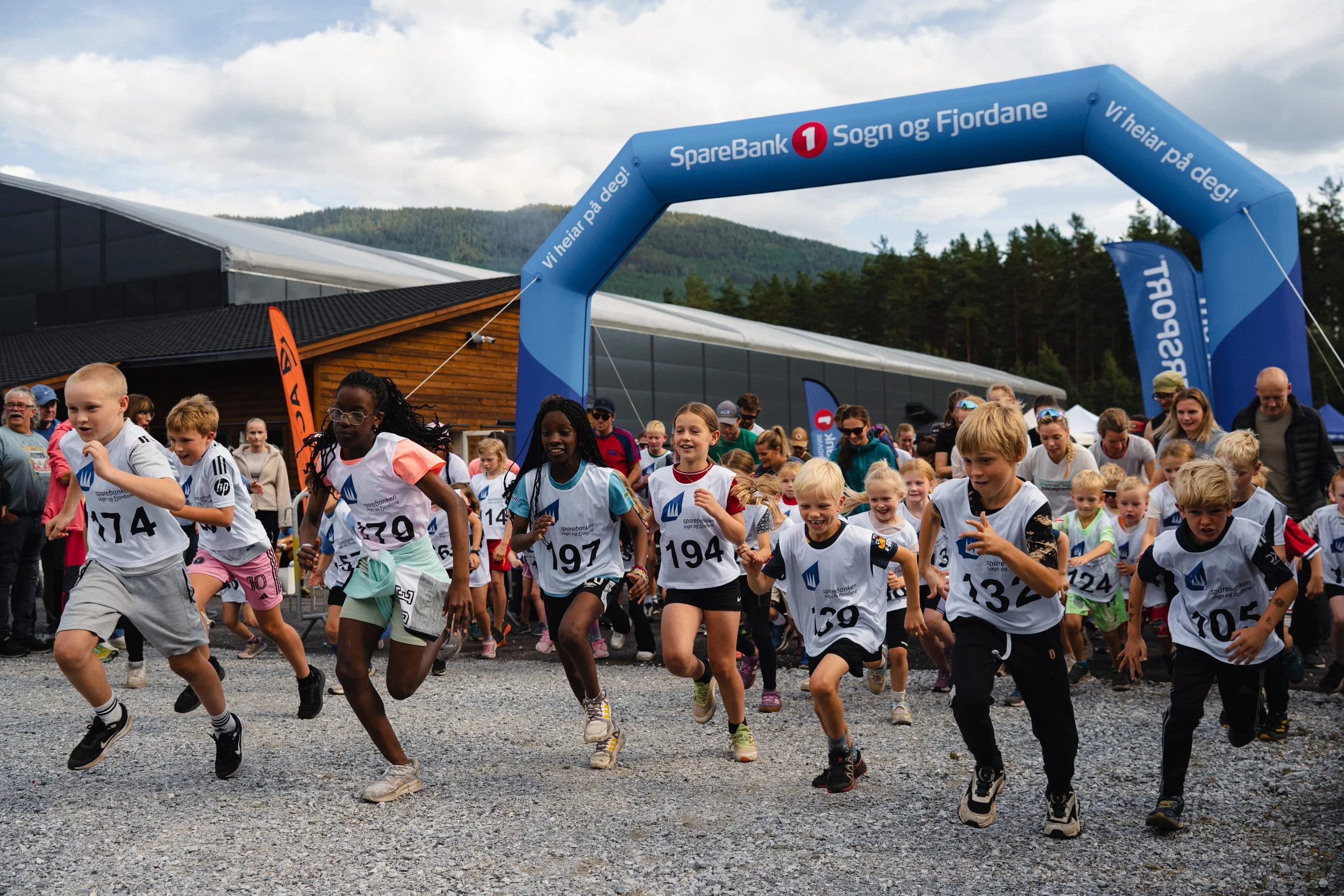 Children participating in a running race at an outdoor event, starting under a blue inflatable archway with sponsors' logos, with spectators and trees in the background.