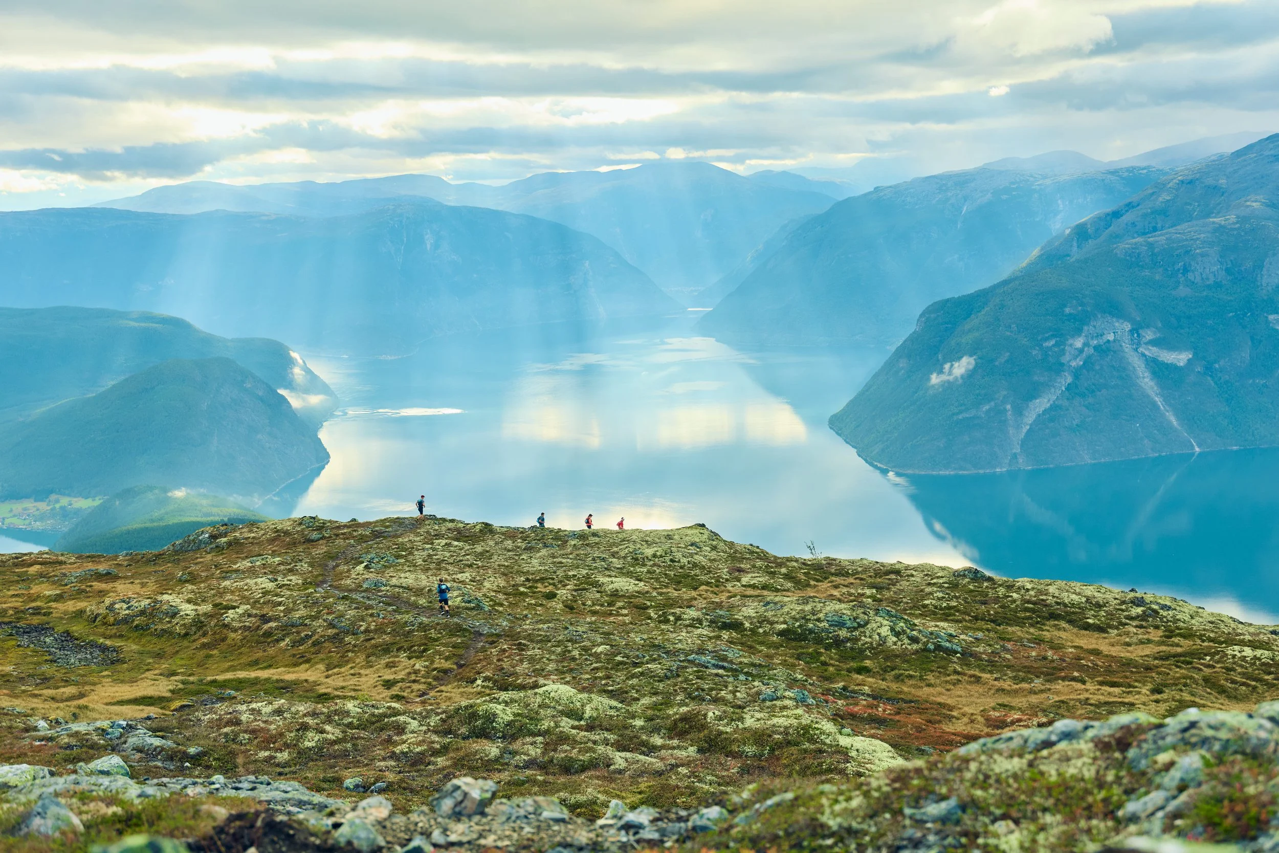 Hikers walking through a grassy, rocky mountain landscape towards a large fjord with steep, green mountains on either side and cloud-covered sky.