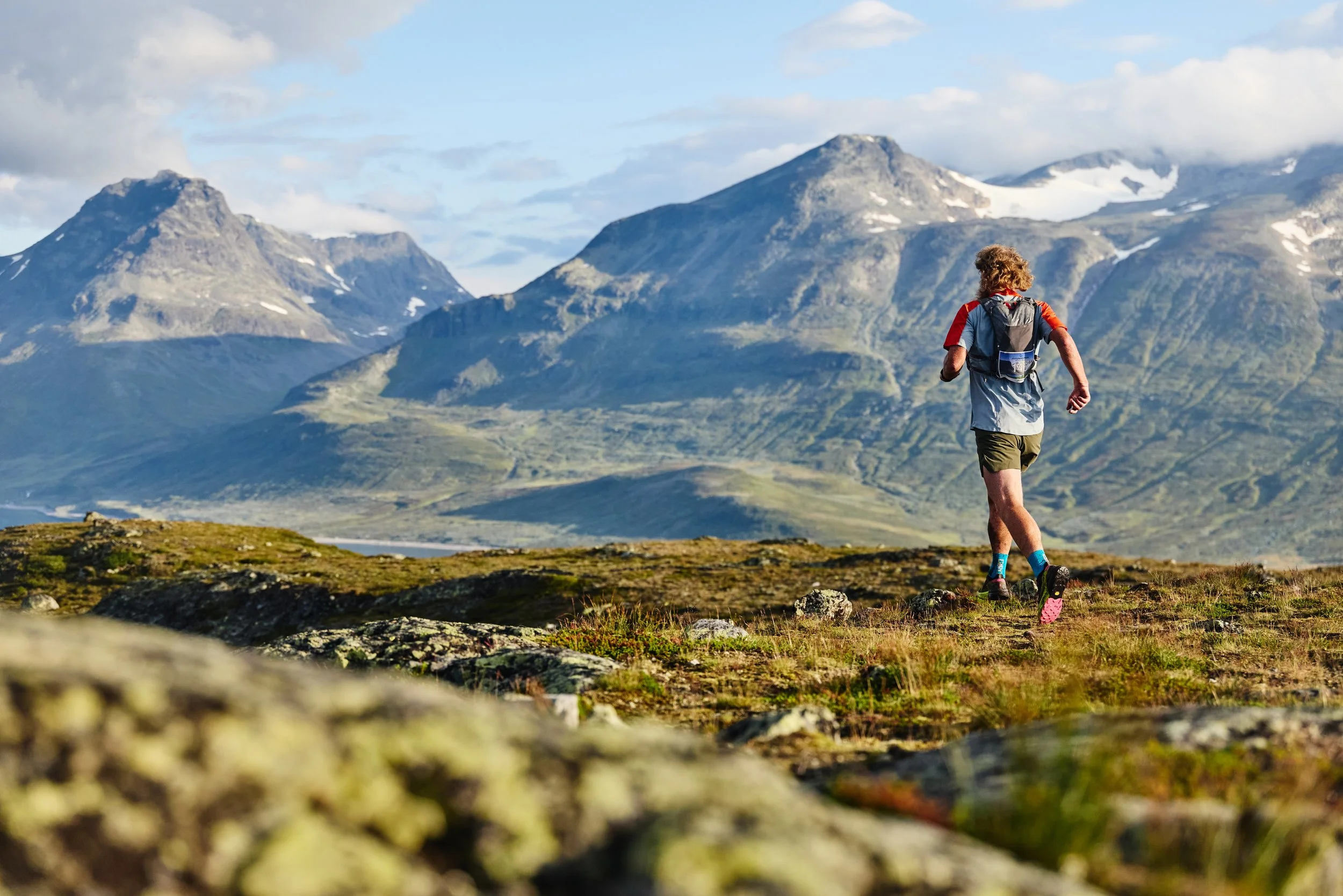 A person with curly hair hiking in a mountainous landscape with green valleys and snow-capped peaks in the background.