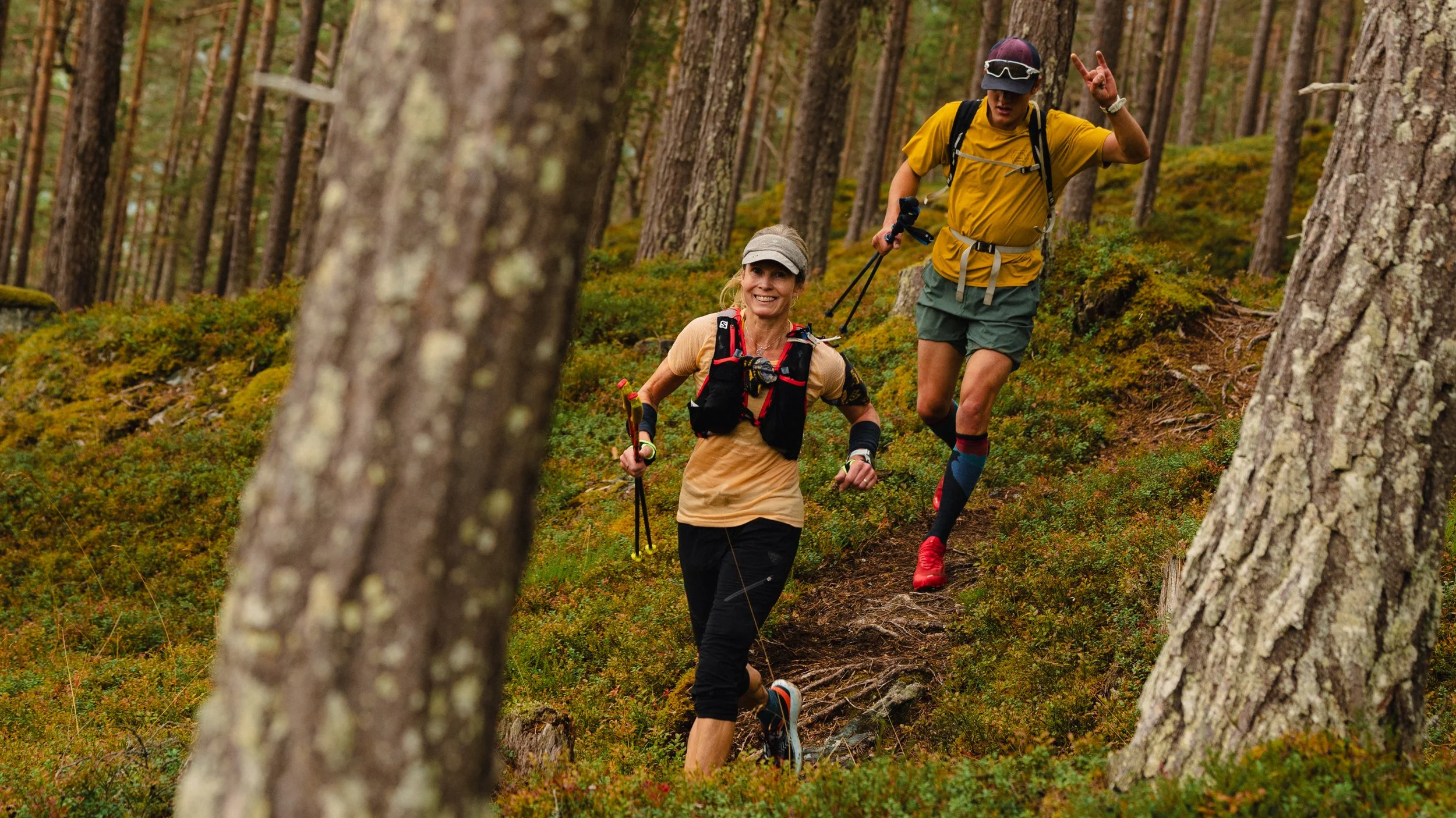 Two runners, a woman and a man, trail running through a forested area with tall trees and mossy ground, smiling and making gestures.
