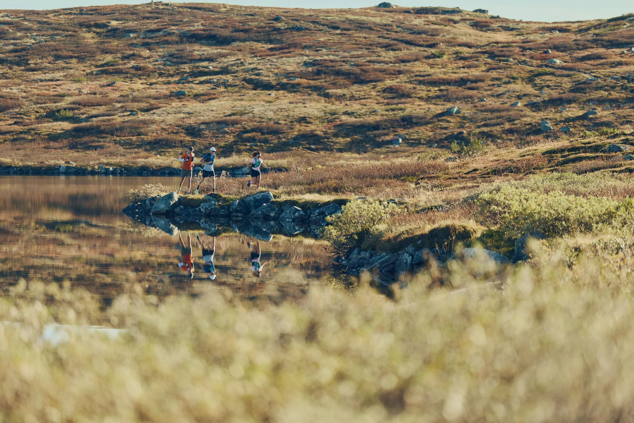 Three people hiking along a rocky path beside a calm lake in a mountainous landscape, with dry grass and shrubs.