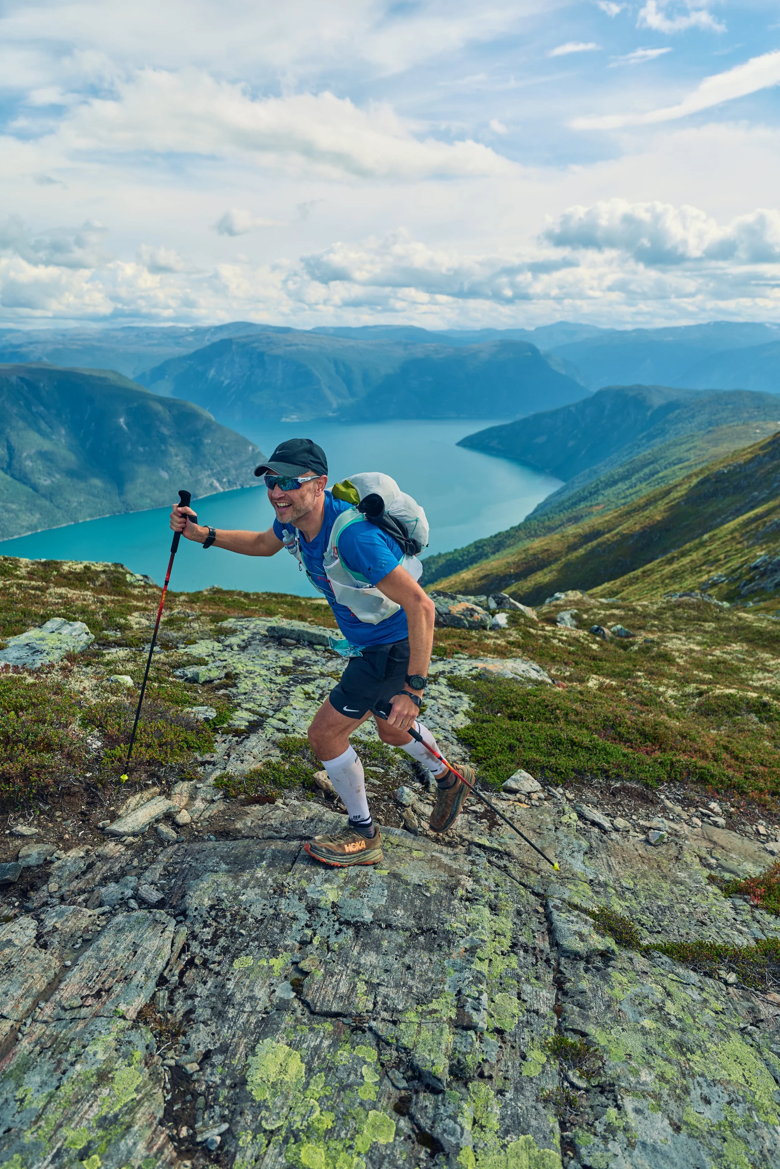 A man hiking uphill on a trail with a scenic lake and mountains in the background under a partly cloudy sky.