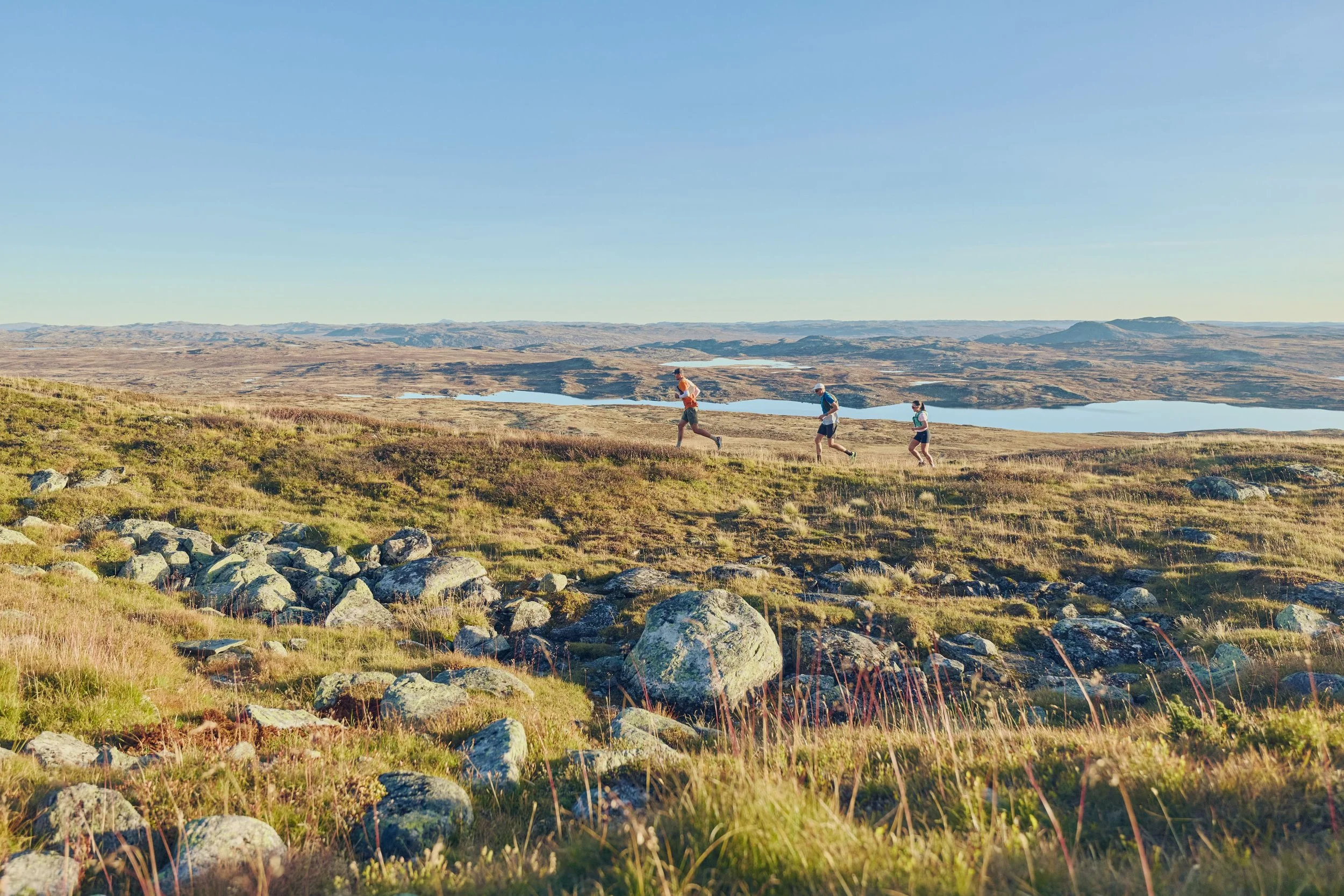Three people trail running on a grassy hillside with rocks, overlooking lakes and rolling hills under a clear blue sky.