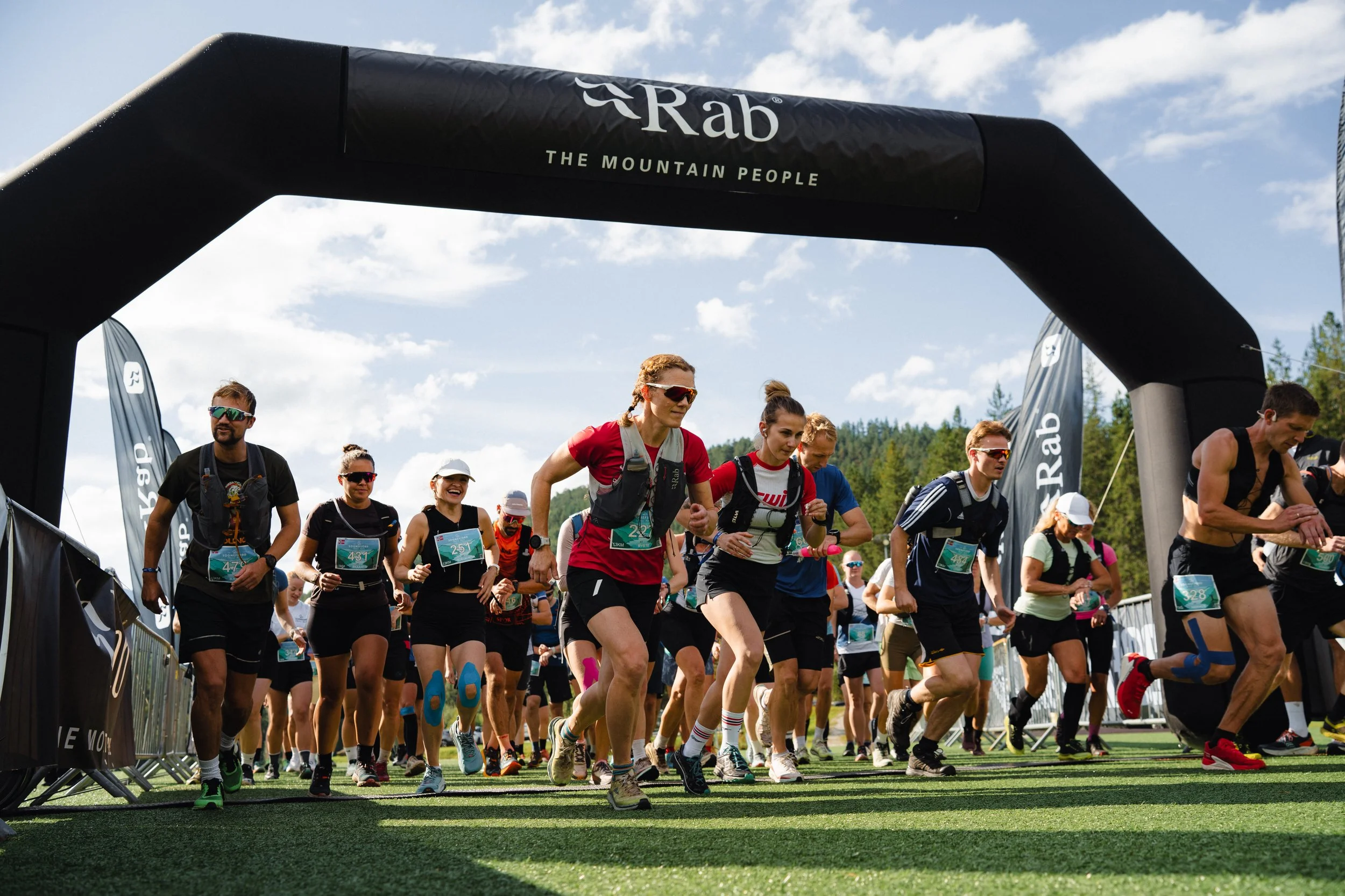 Group of runners starting a race under an archway with 'Rab' branding, on a sunny day with trees in the background.