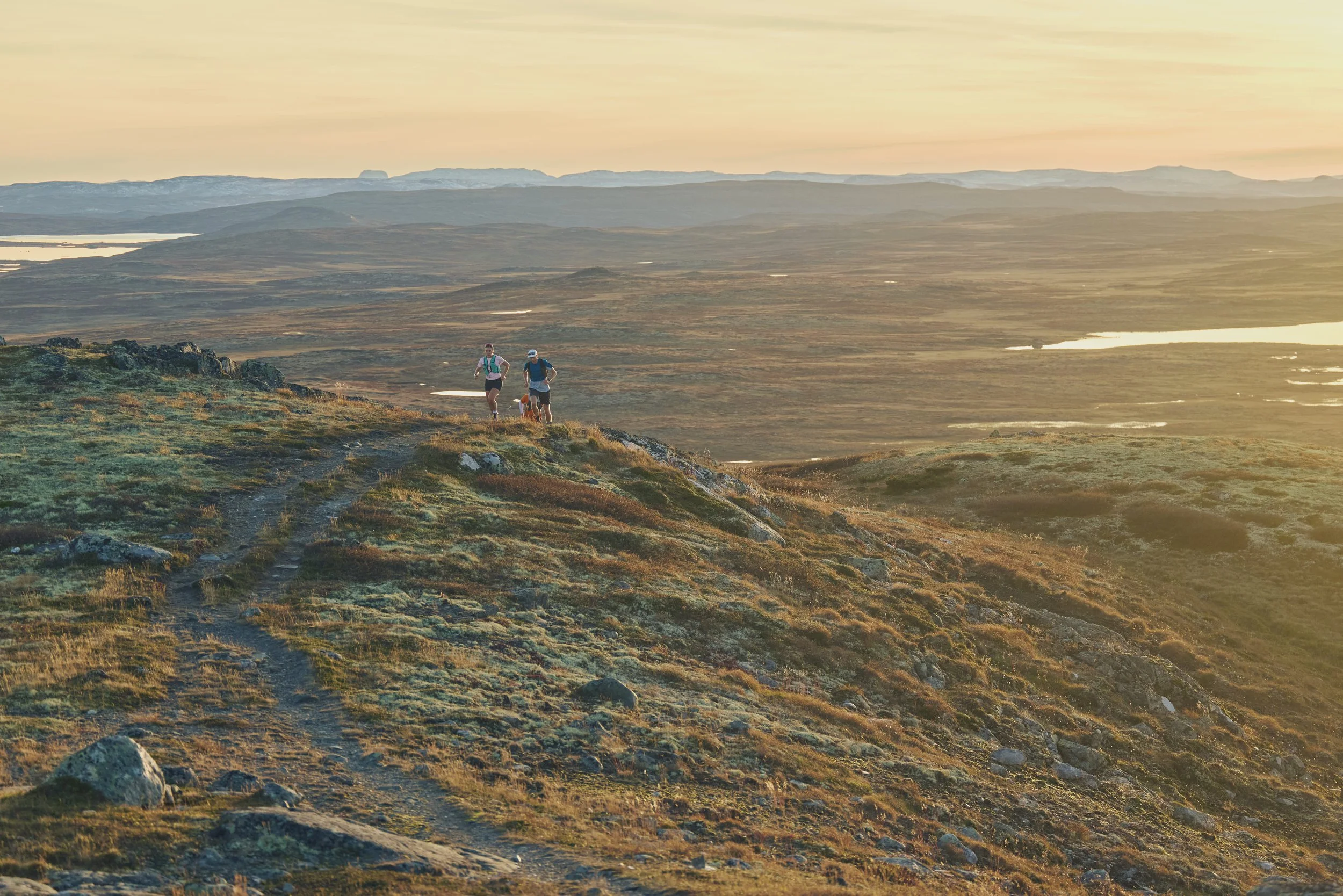 Two hikers walking along a trail on a grassy hillside during sunset in a vast, open landscape with distant mountains and water bodies.